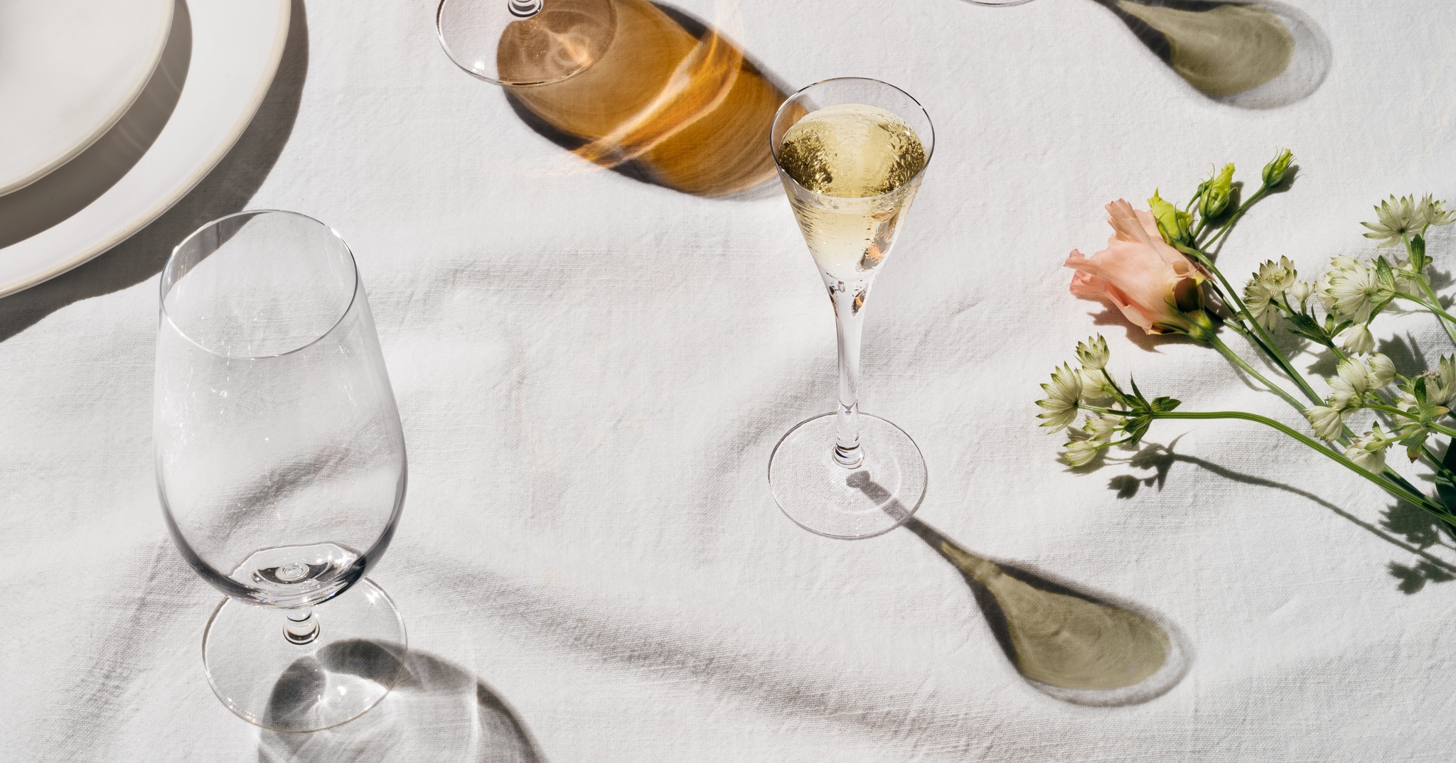 Elegant borddekking med øl, musserende vin, poteter og blomster på hvit duk i sollys.