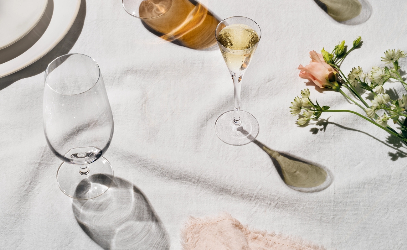 Somrig dukning med öl, mousserande vin, potatis och blommor på vit linneduk i mjukt solljus.