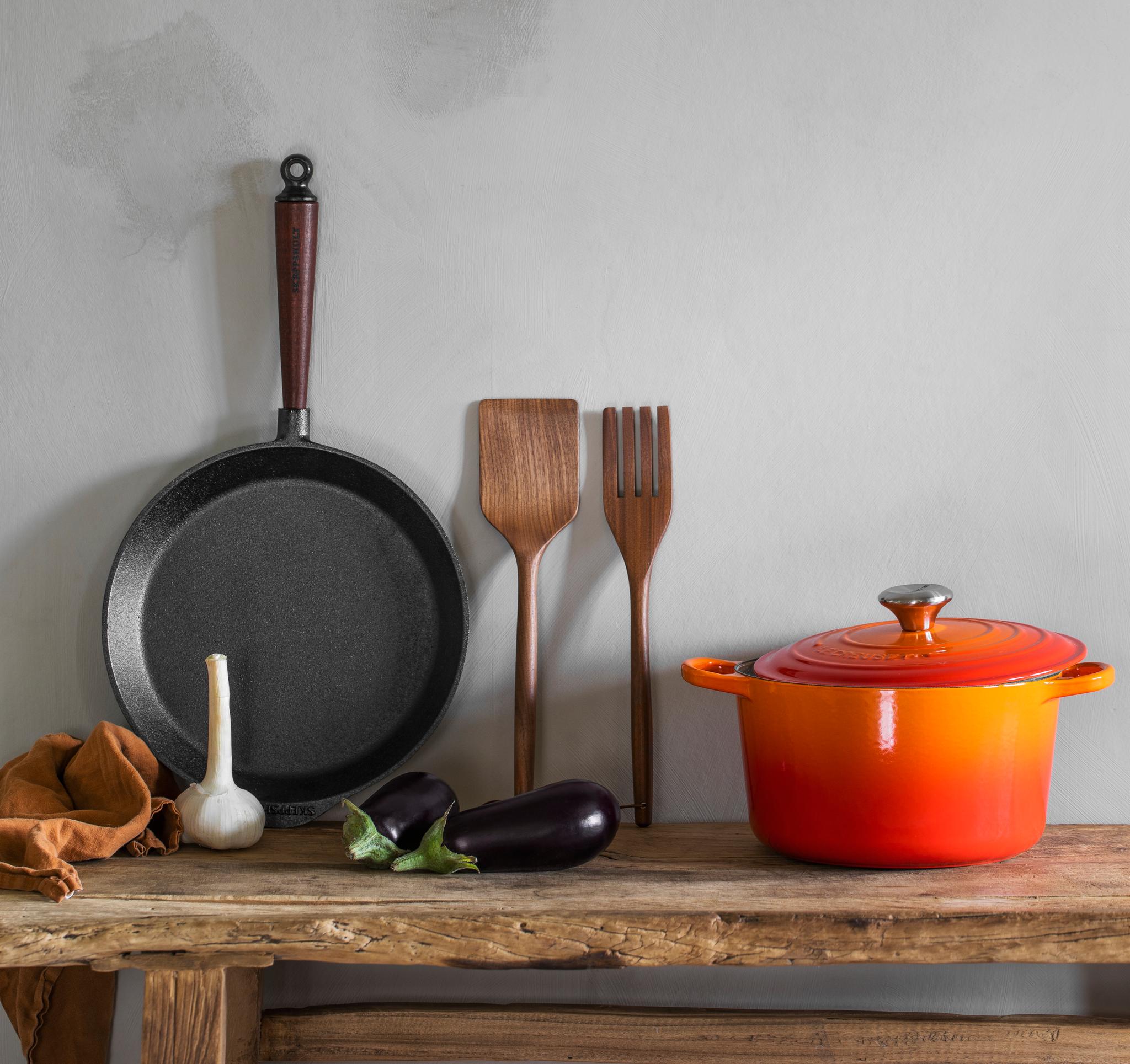 Gjutjärnspanna, träredskap och orange gryta med auberginer och vitlök på ett rustikt träbord.