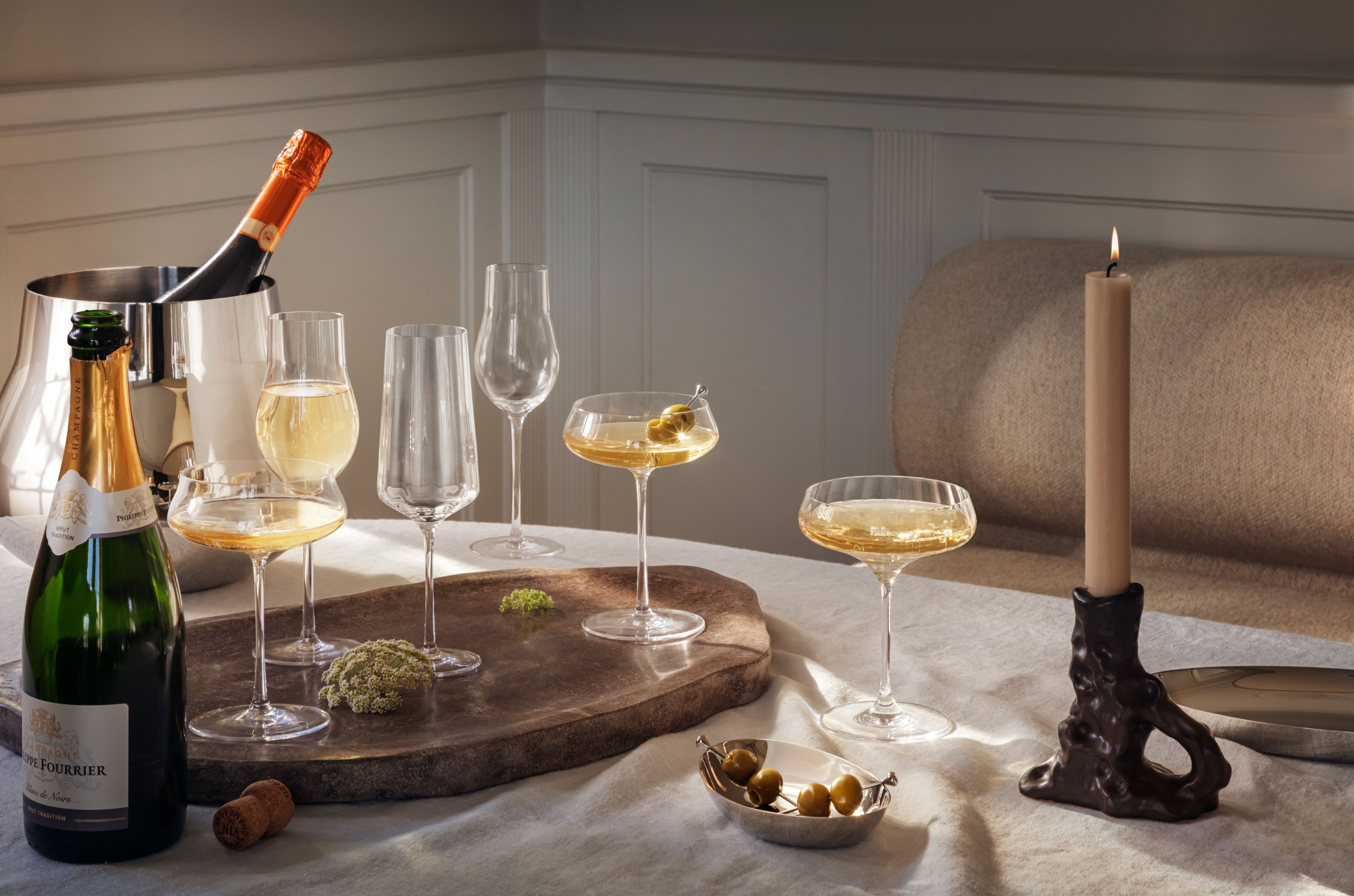 Elegant dukning med champagneglas, flaskor, gröna oliver och tänd ljusstake på ett stilfullt bord.