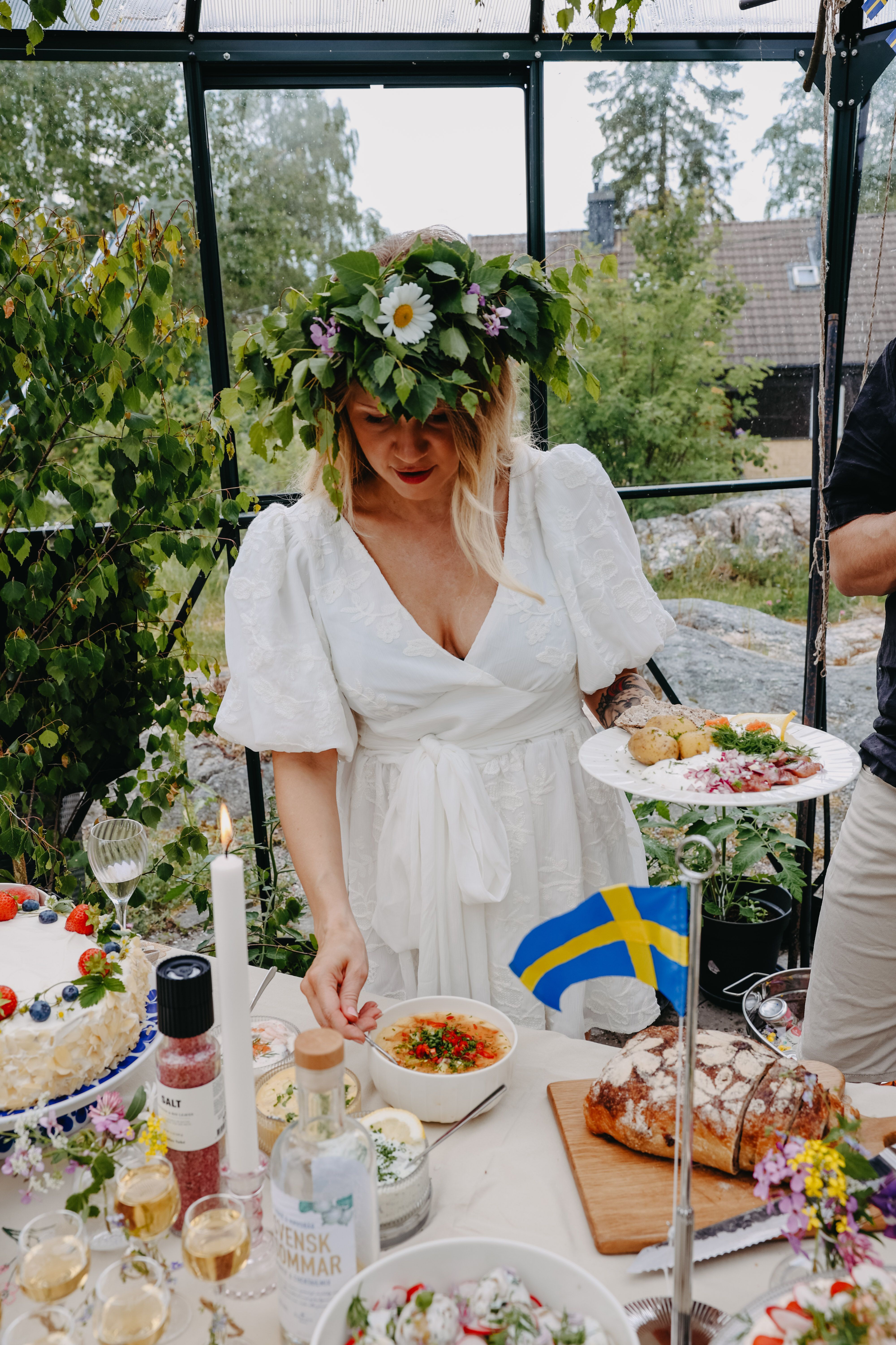 Kvinna i vit klänning med midsommarkrans står vid ett sommarbord dukat med traditionell svensk mat och liten svensk flagga.