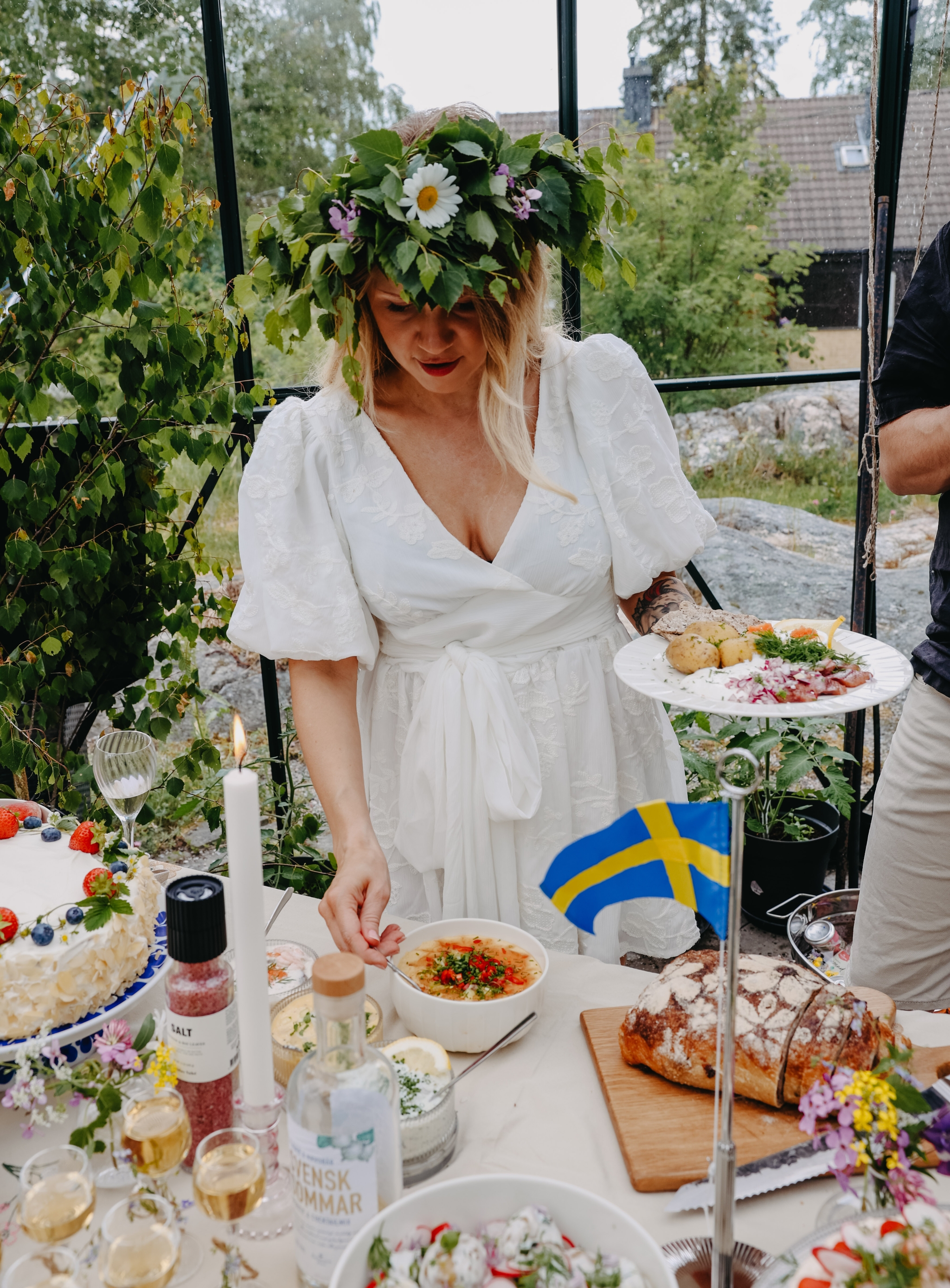 Kvinna i vit klänning med midsommarkrans står vid ett sommarbord dukat med traditionell svensk mat och liten svensk flagga.
