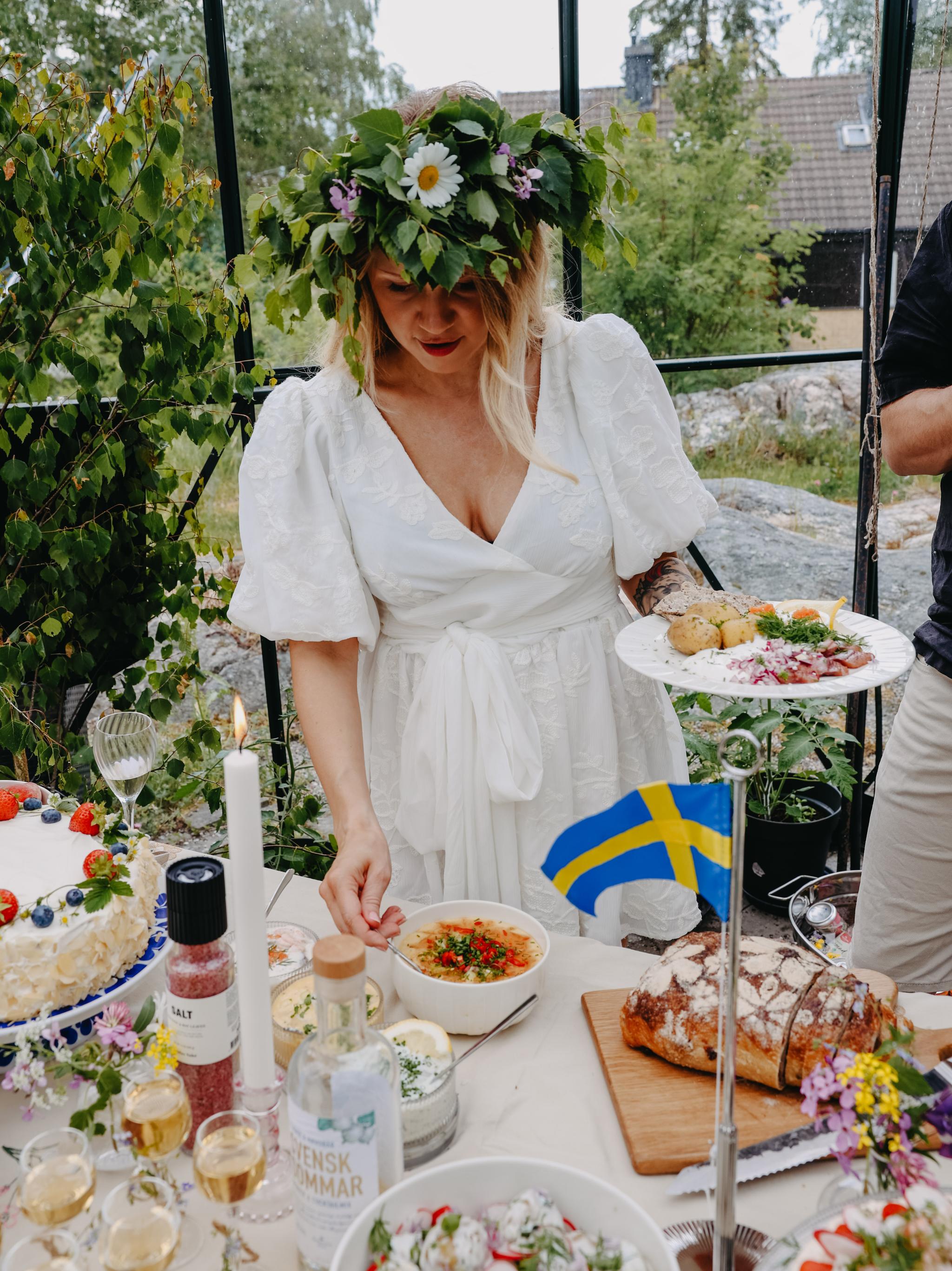 Kvinna i vit klänning med midsommarkrans står vid ett sommarbord dukat med traditionell svensk mat och liten svensk flagga.