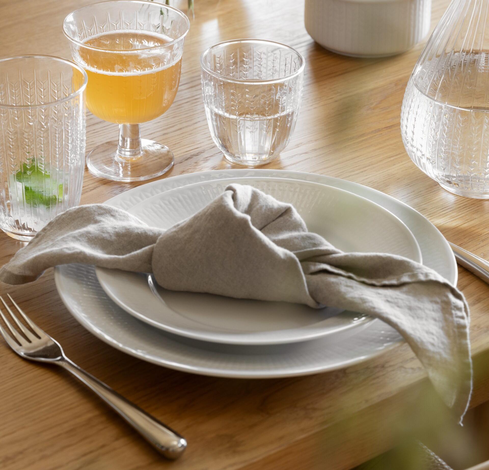 A dining table set with white plates, a grey linen napkin, a fork, and textured glasses holding an amber drink, water, and a garnished beverage on a wooden surface.