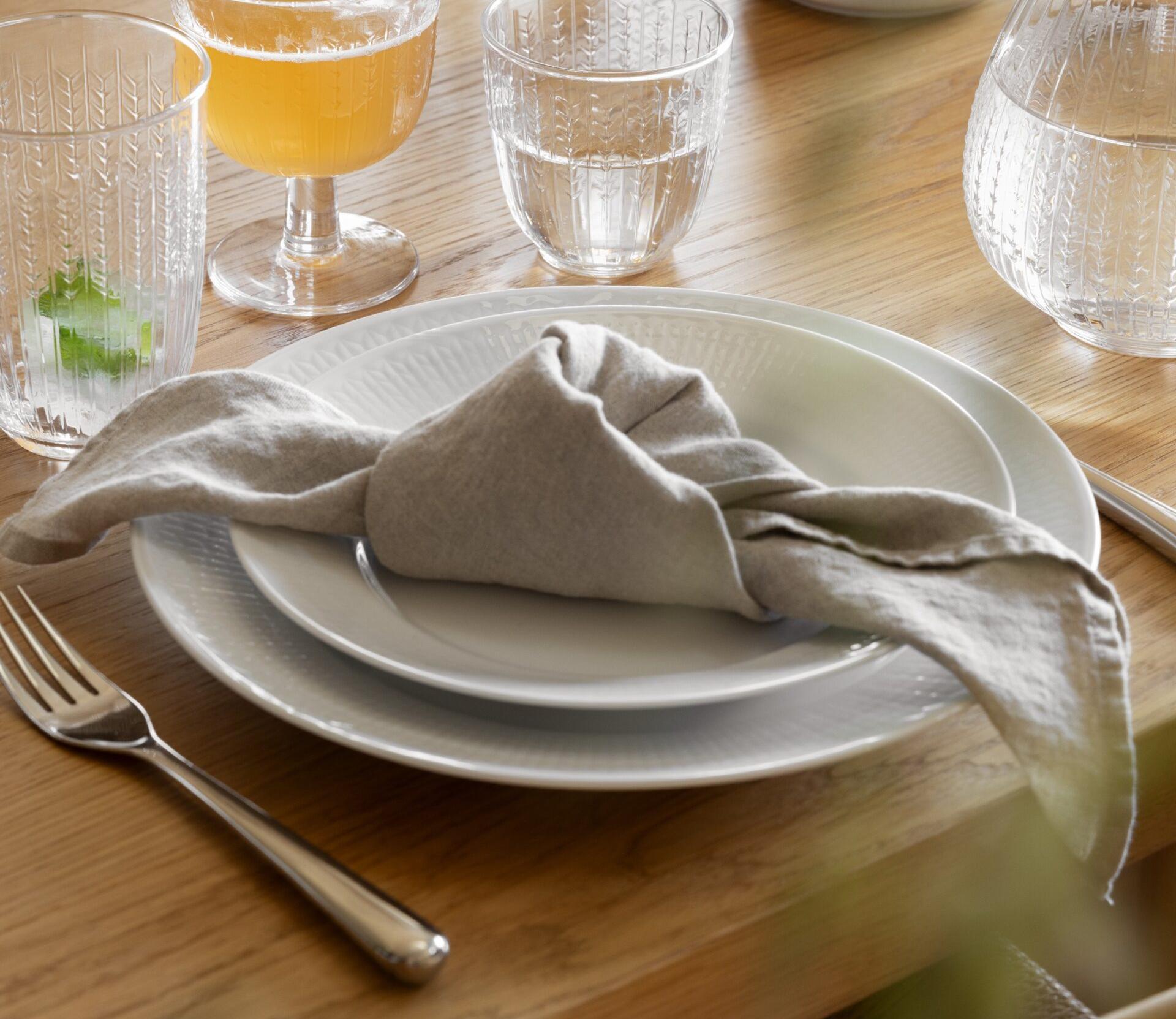 Ruokapöytä, jossa valkoiset lautaset, solmittu harmaa pellavalautasliina, hopeinen haarukka ja kuviolliset lasit juomineen puupöydällä.