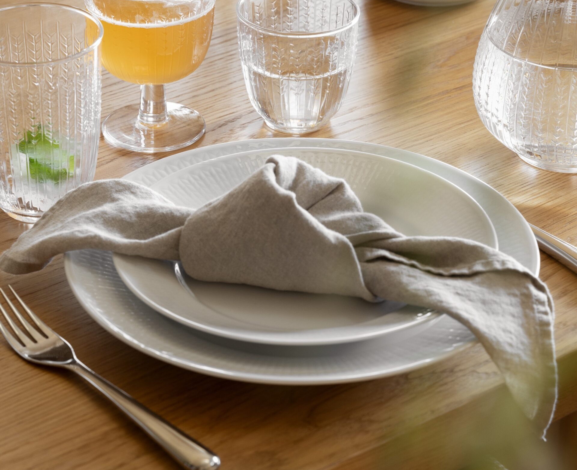 Ruokapöytä, jossa valkoiset lautaset, solmittu harmaa pellavalautasliina, hopeinen haarukka ja kuviolliset lasit juomineen puupöydällä.