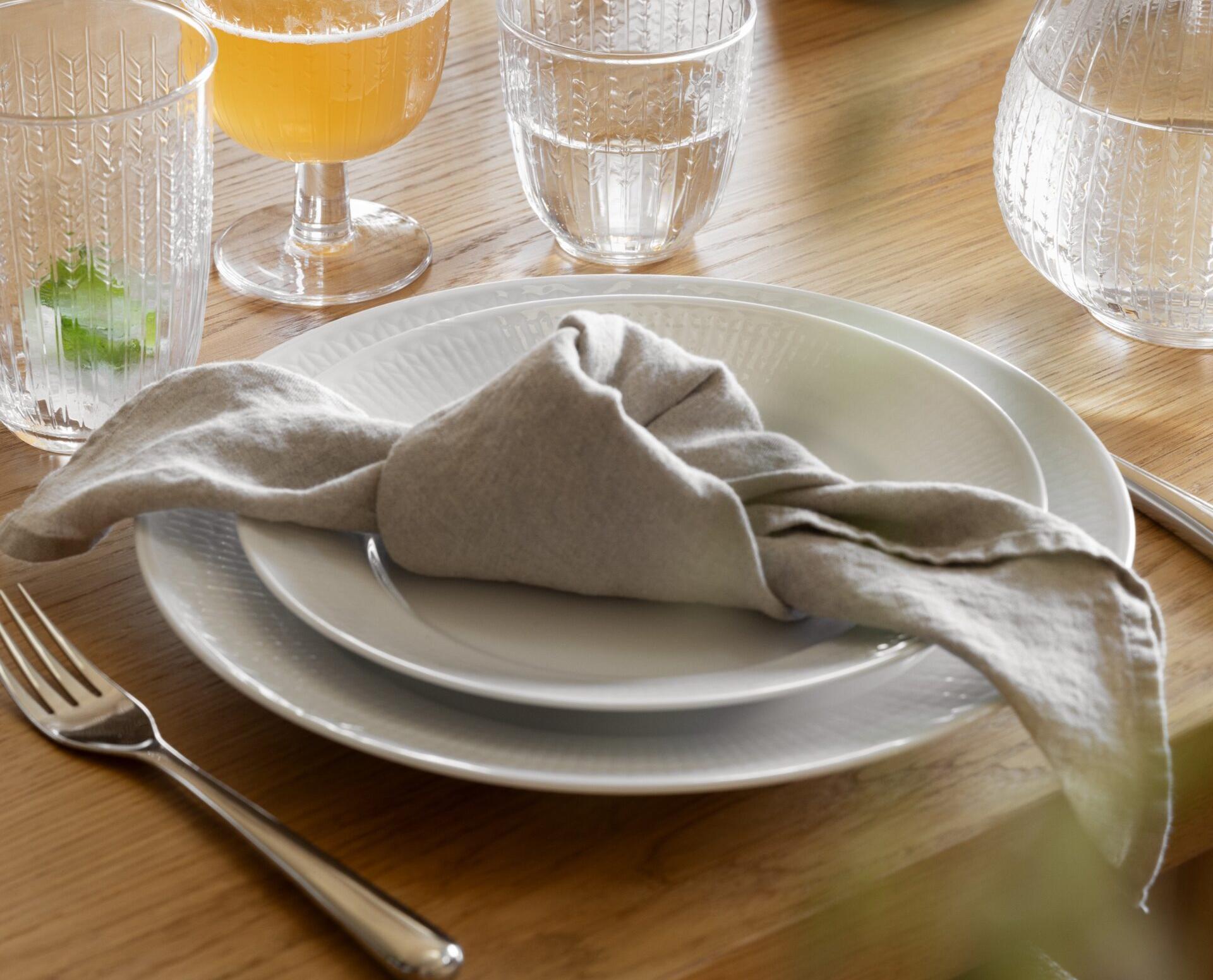 En dukning med vita strukturerade tallrikar, en beige linneservett och två mönstrade glas (ett med en bärnstensfärgad dryck, ett med vatten) på ett ljust träbord. Gröna vetestjälkar ligger i en vas i bakgrunden.
