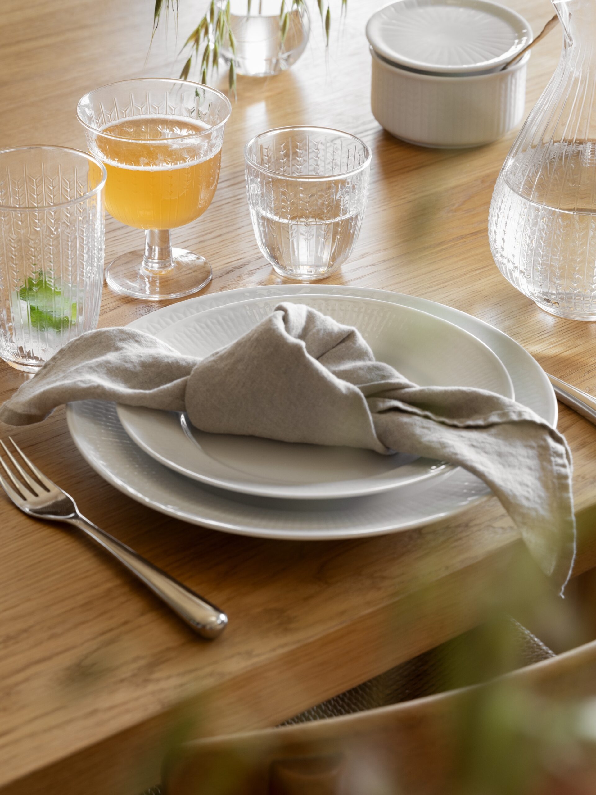A dining table set with white plates, a grey linen napkin, and various drinks in patterned glassware on a light wooden surface.