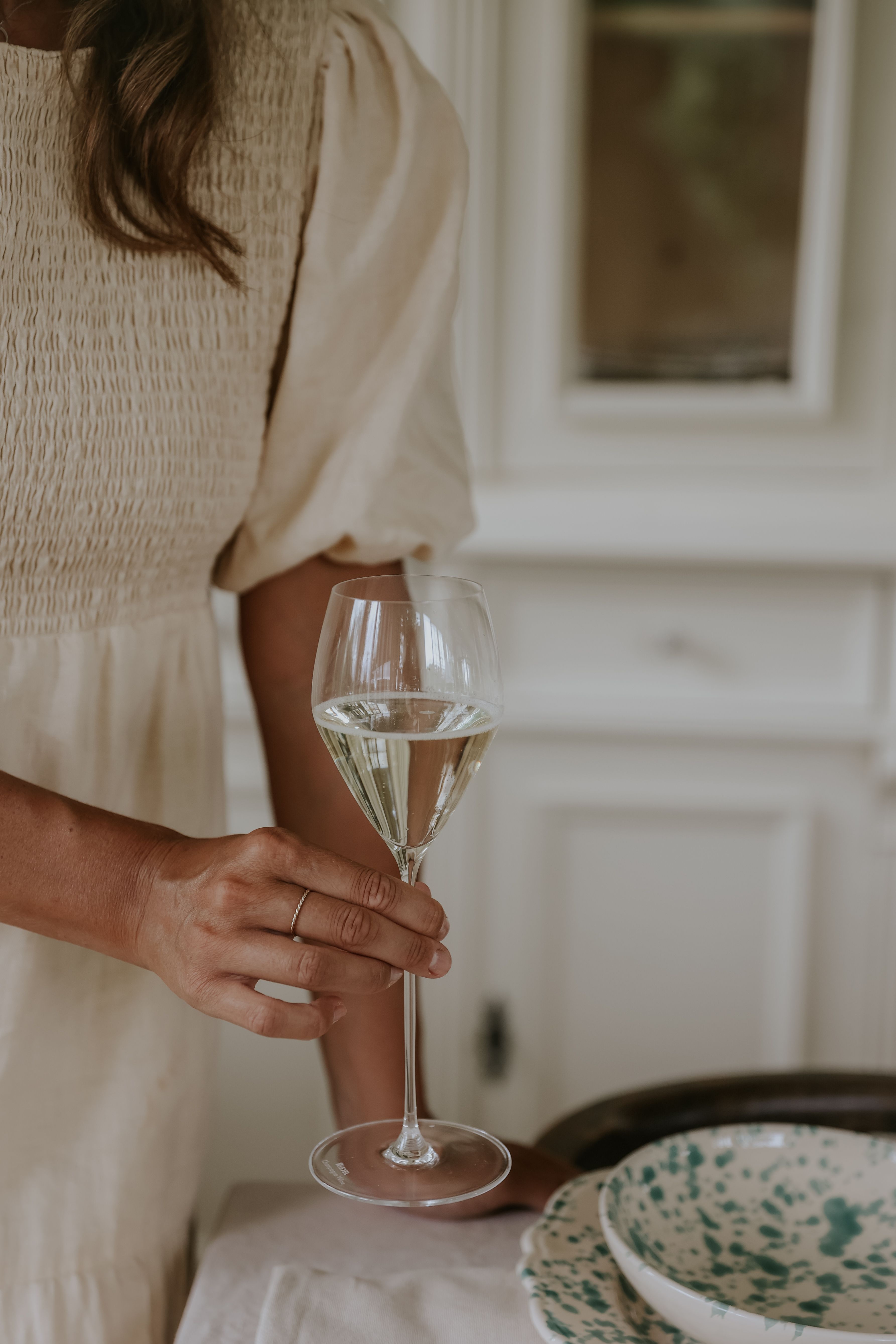 Kermanväriseen rypytettyyn mekkoon pukeutunut henkilö pitelee lasillista valkoviiniä.