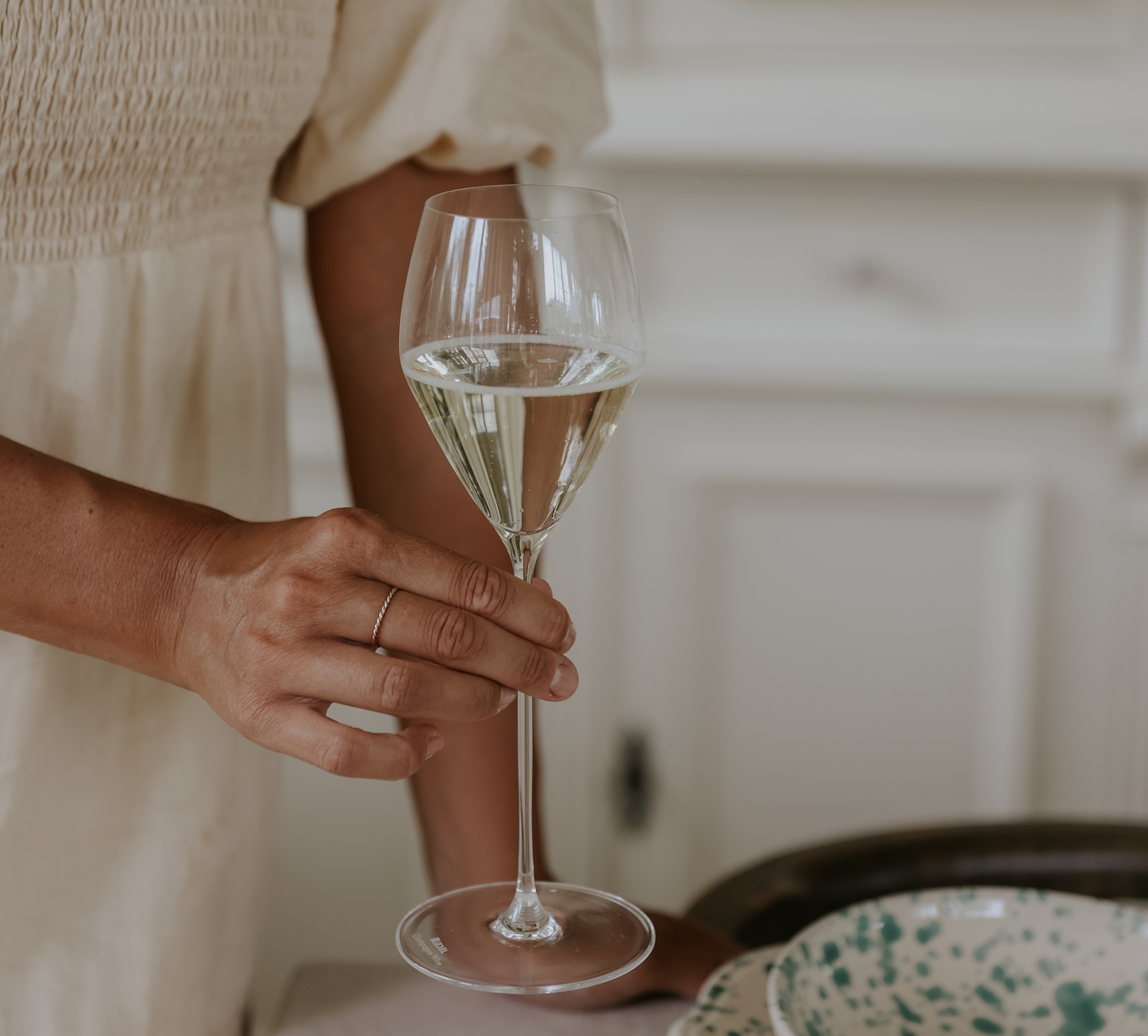 Person i ljus klänning håller ett glas vitt vin vid ett dukat bord i ett ljust kök.