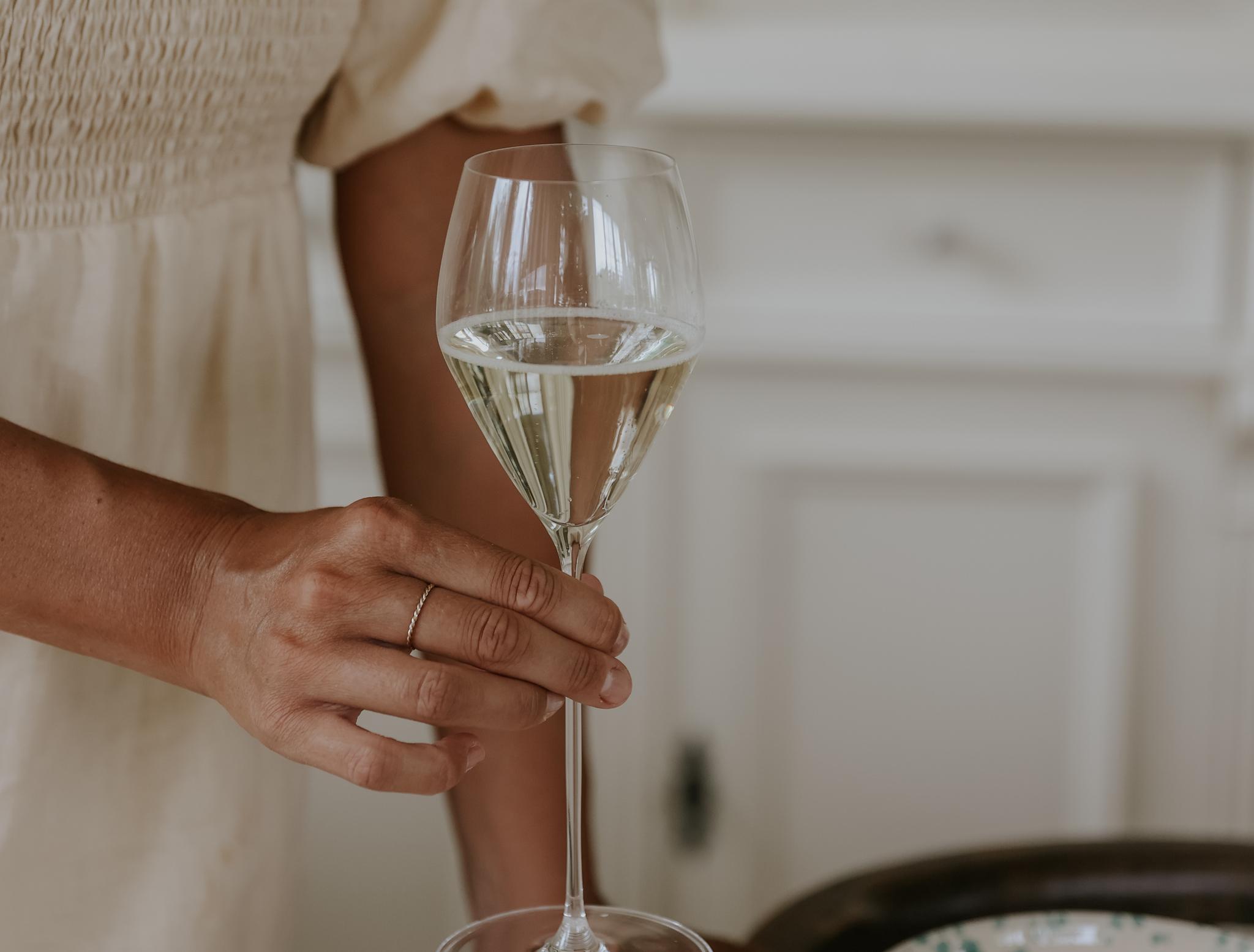 Person i ljus klänning håller ett glas vitt vin vid ett dukat bord i ett ljust kök.