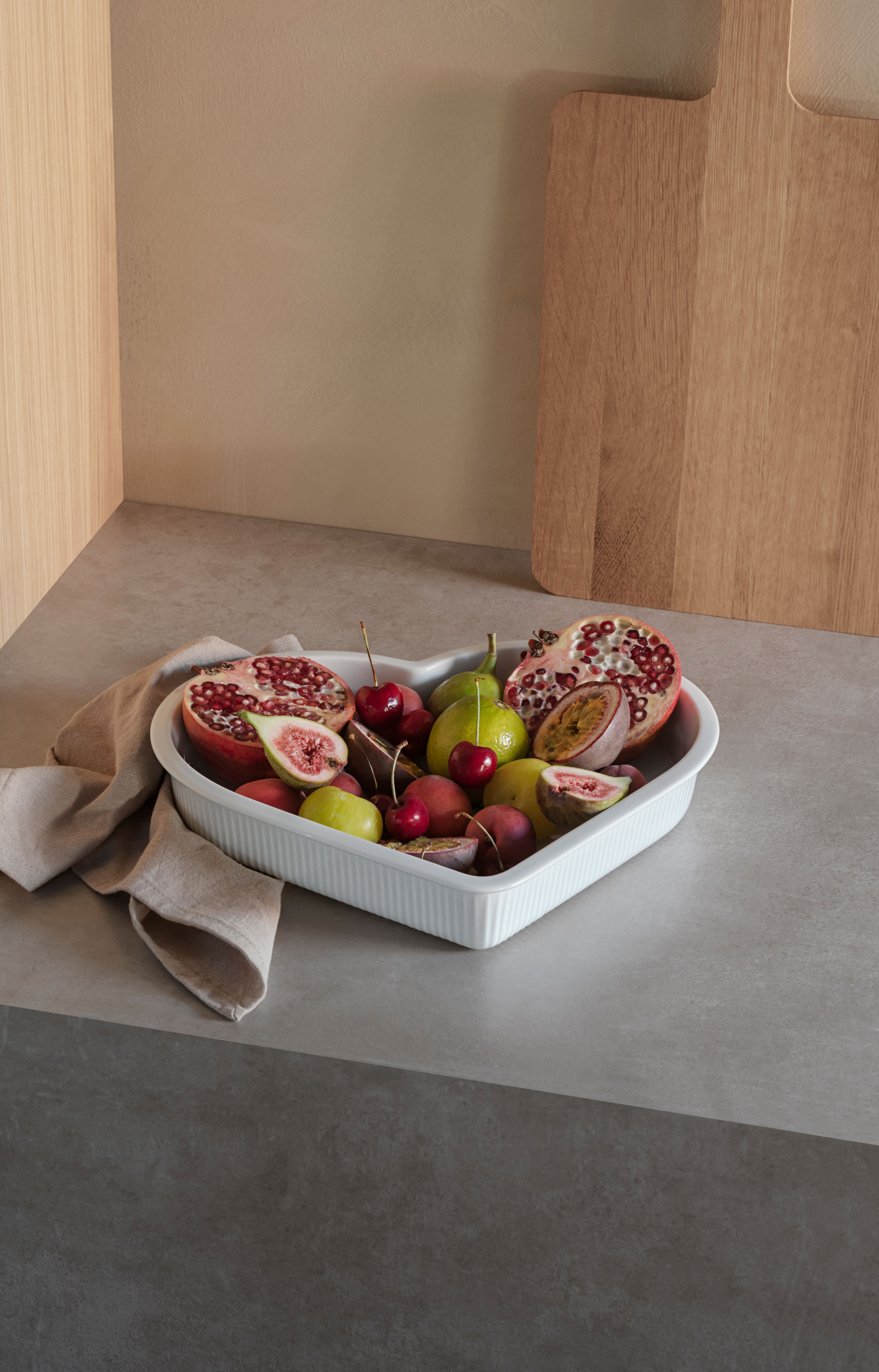 En vit hjärtformad skål fylld med skurna granatäpplen, fikon, passionsfrukt, körsbär och andra färgglada frukter, på en bänk med en linneduk.