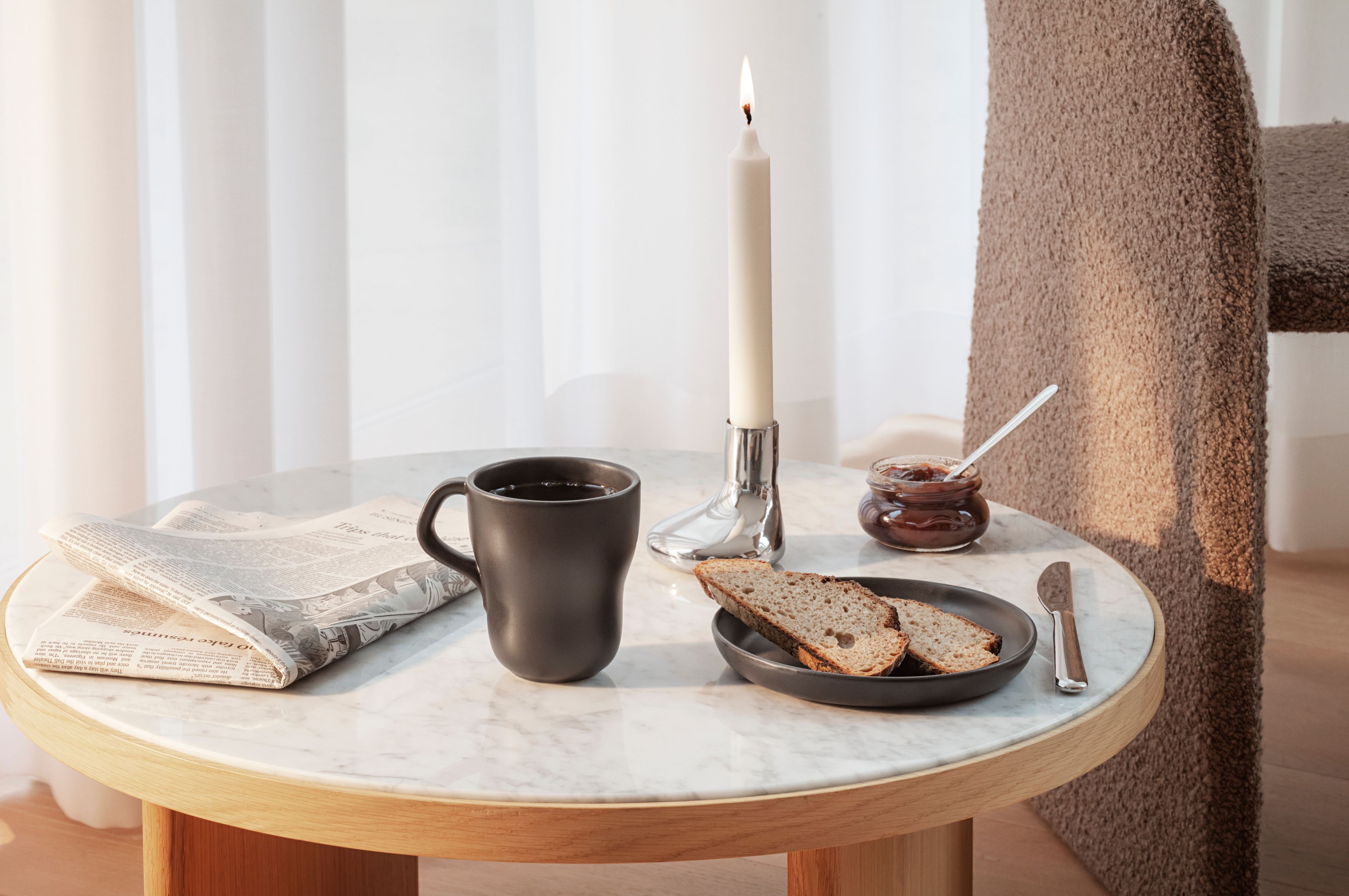  En mysig frukostscen med en svart mugg, tidning, tänt ljus, bröd och sylt på ett marmorbord