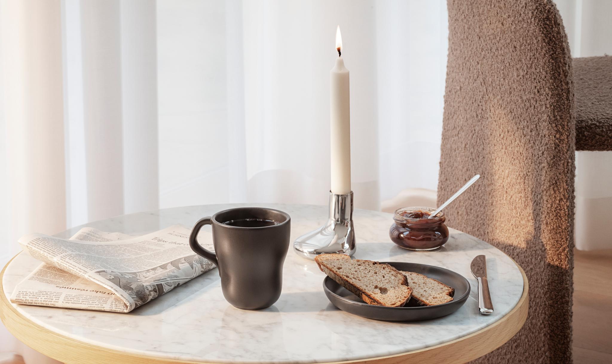 Ljusstake från eva solo står tänt på ett bord tillsammans med en kaffemugg, samt en tidning