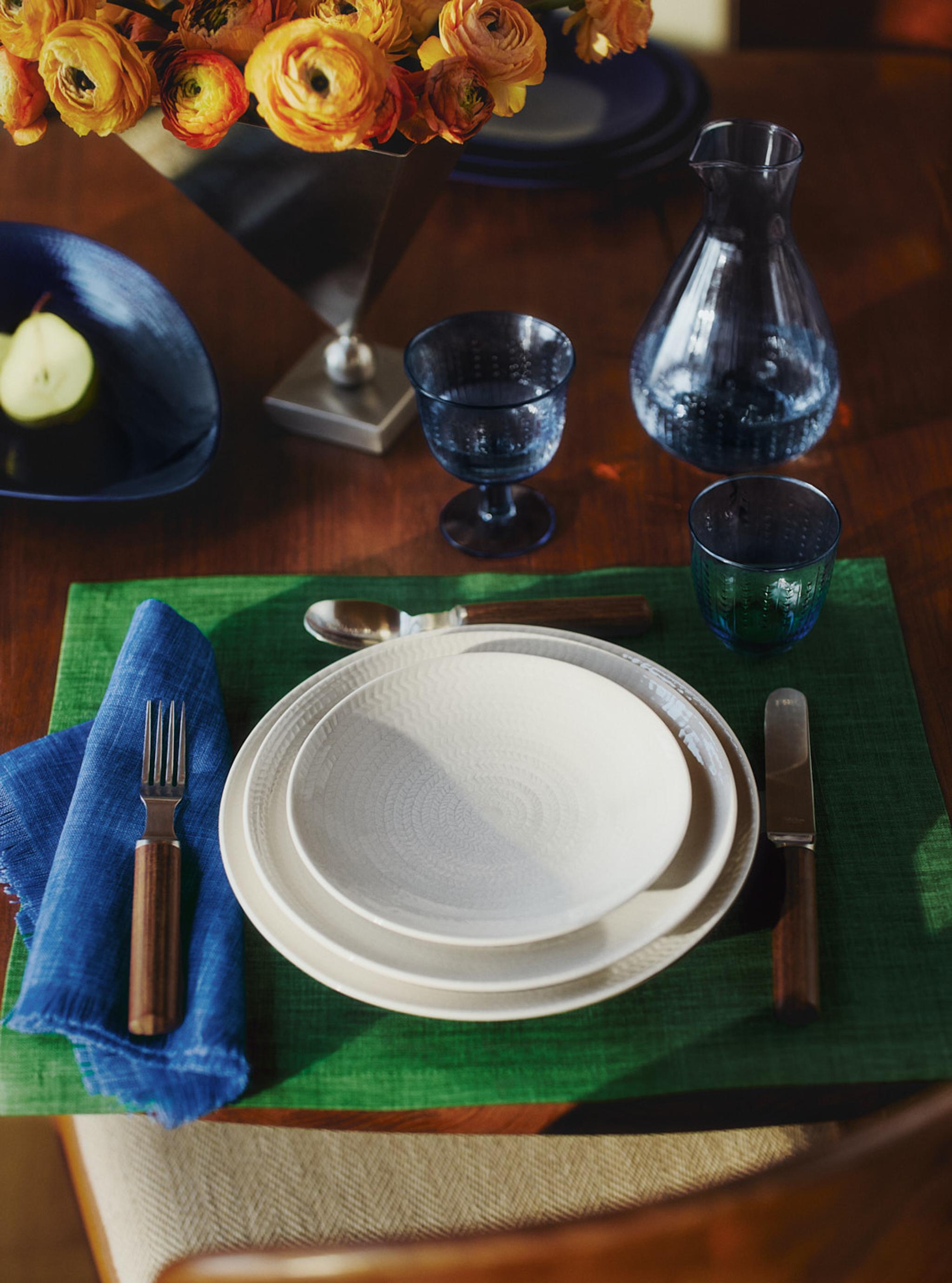 Ett dukat bord med vita strukturerade tallrikar, en grön bordsunderlägg, blått glas, en vas med orange ranunkel och en blå skål med ett päron.