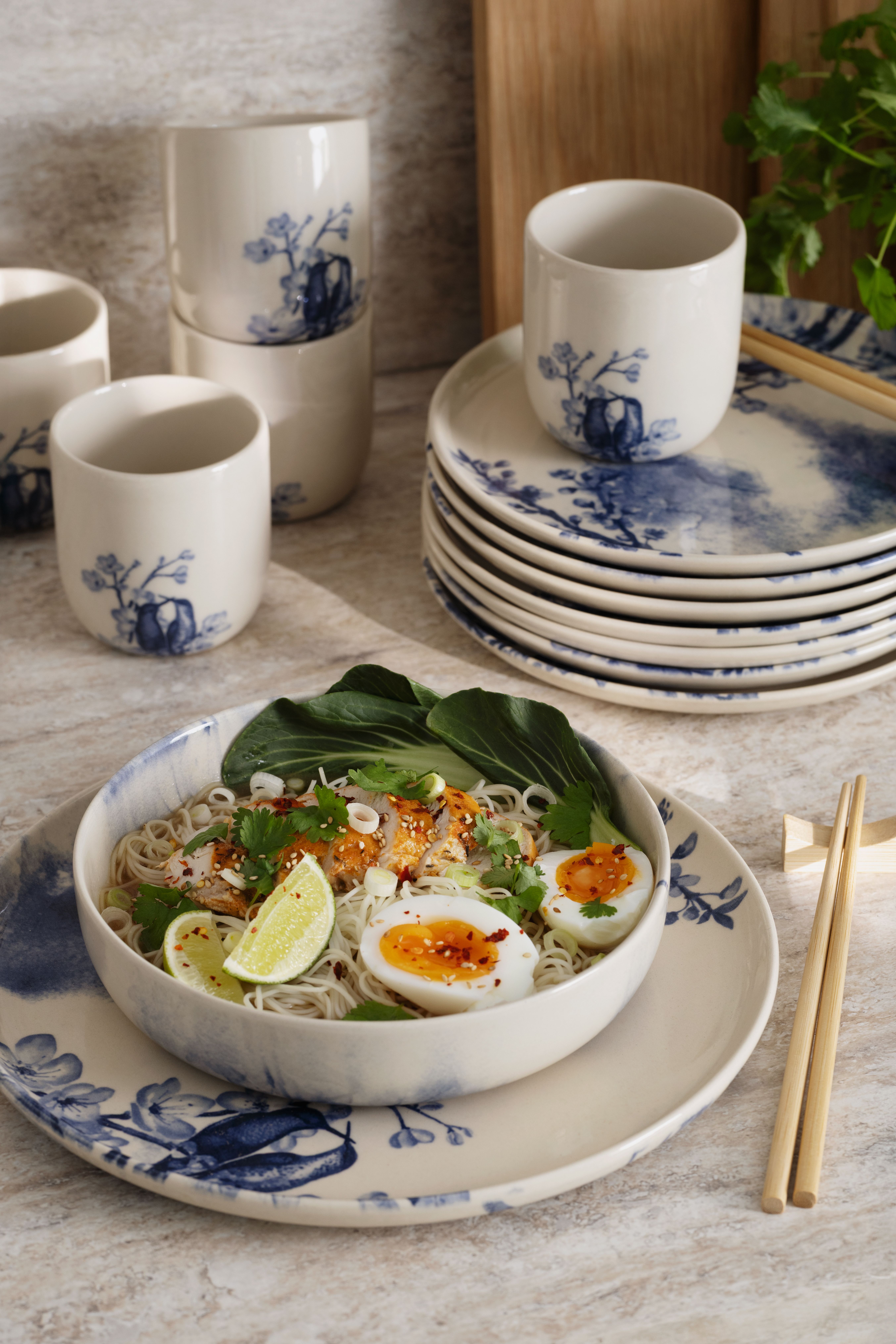 En skål ramen med kyckling, ägg och lime på en blåvitmönstrad keramisk tallrik, med högar av matchande porslin i bakgrunden.
