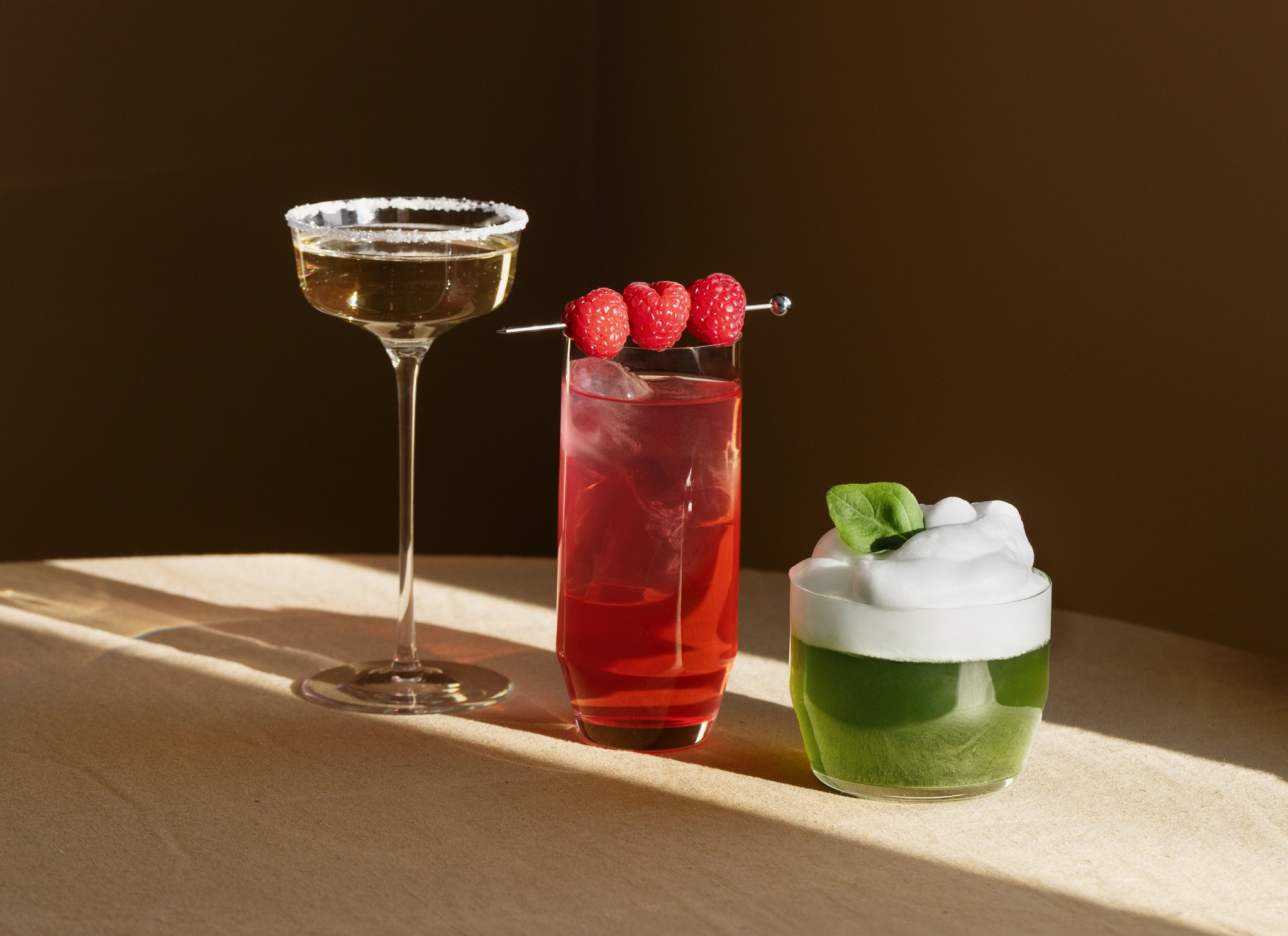 Tre färgglada cocktails – en gyllene, en röd med hallon och en grön med skum – står upplysta på ett bord mot en mörk bakgrund.