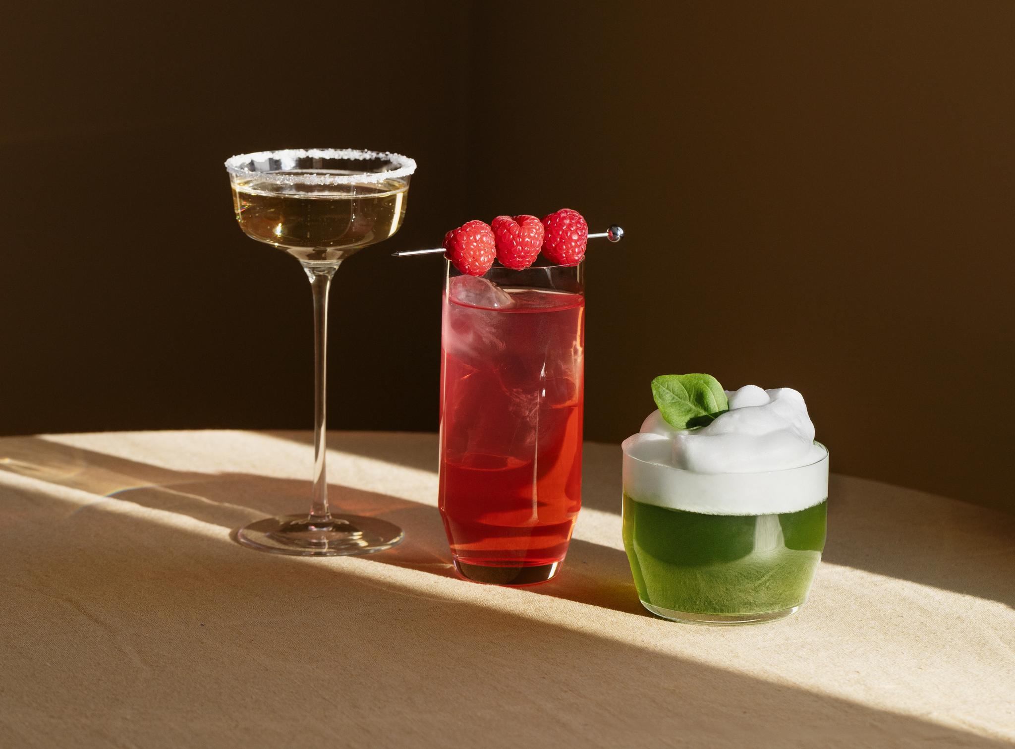 Tre färgglada cocktails står uppställda på ett bord i varmt solljus, en gyllene drink, en röd hallondrink och en grön drink med skum och basilika.