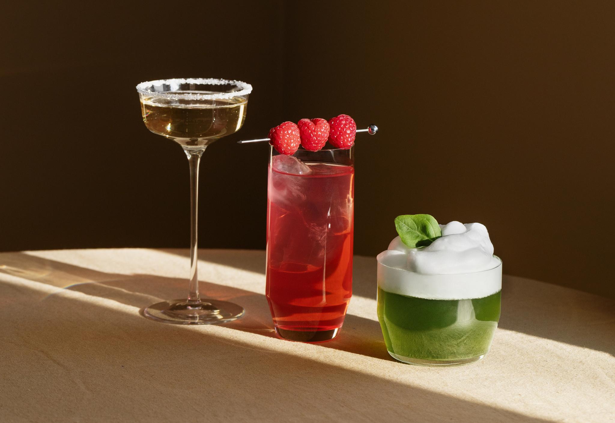 Tre färgglada cocktails – en mousserande, en röd med hallon och en grön med skum – står upplysta på ett bord mot en mörk bakgrund.
