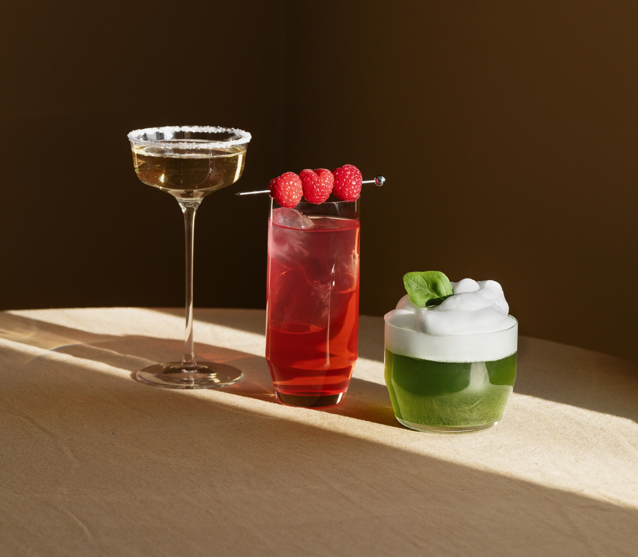 Tre färgglada cocktails på ett bord i varmt solljus, en gyllene drink, en röd hallondrink och en grön drink med skum och basilika.