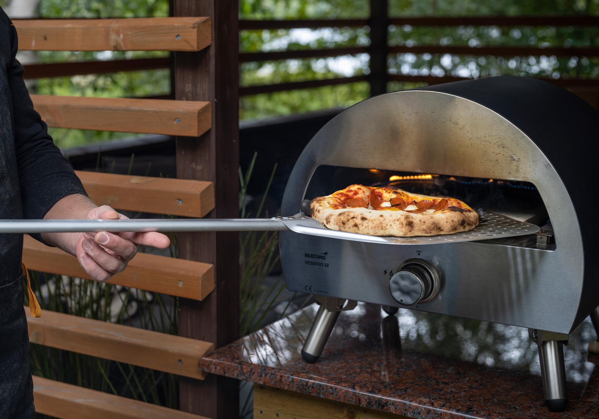 En mann skyver en pizza med en pizzaspade inn i en utendørs gassdrevet pizzaovn