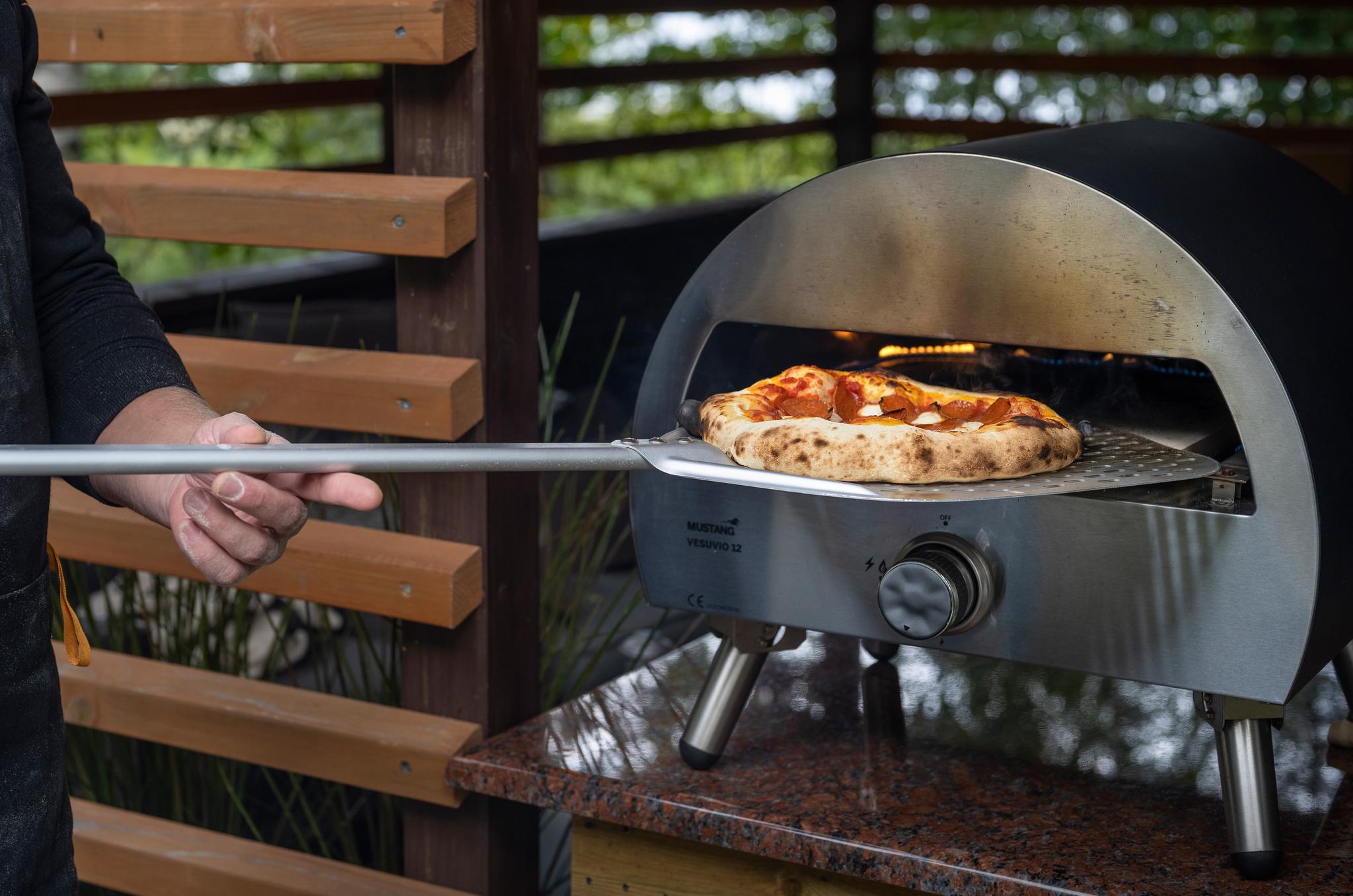En man skjuter in en pizza med en pizzaspade i en utomhus gasdriven pizzaugn.