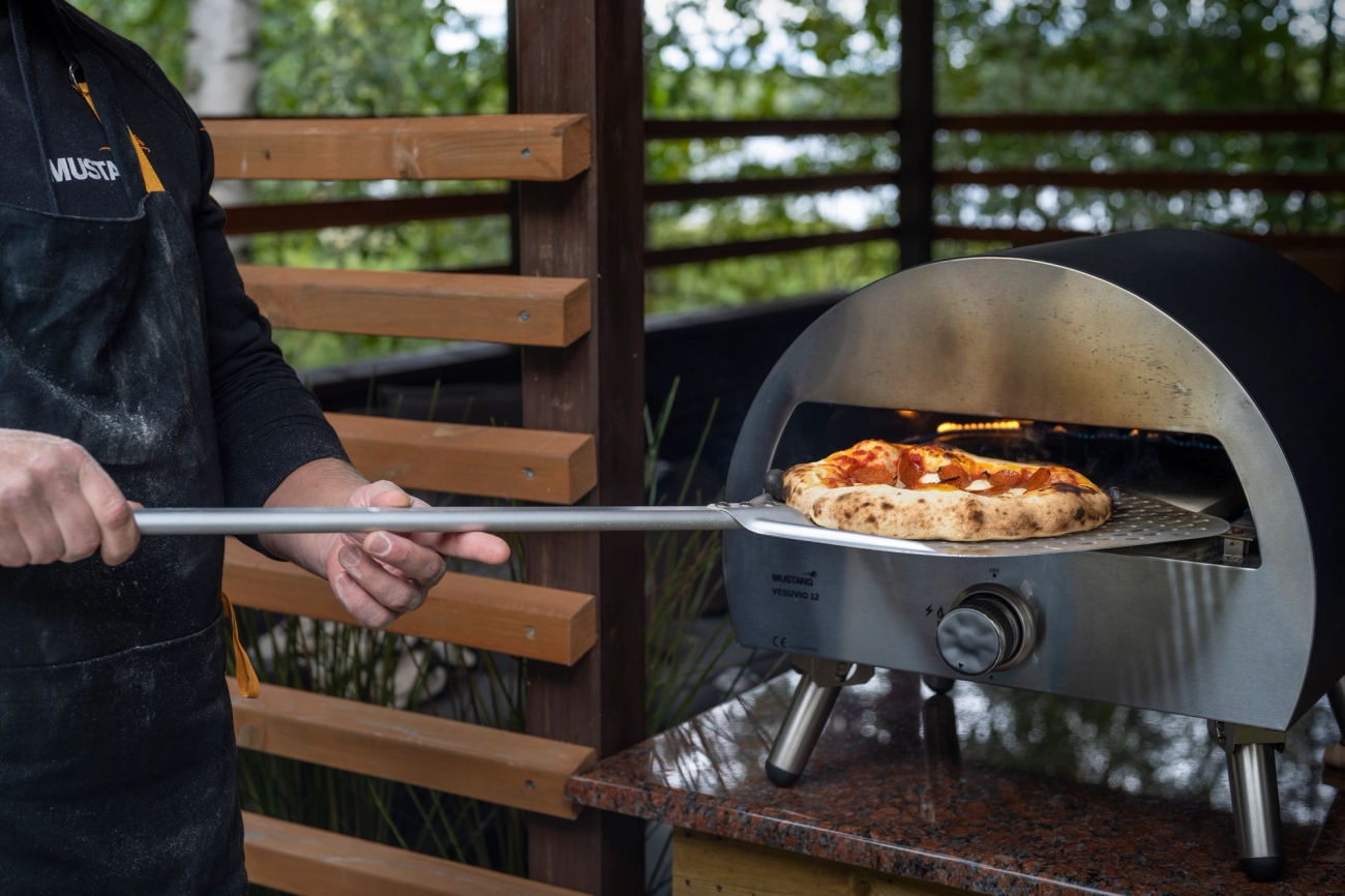 Henkilö paistaa napolilaista pizzaa teräksisessä ulkouunissa puutarhassa.