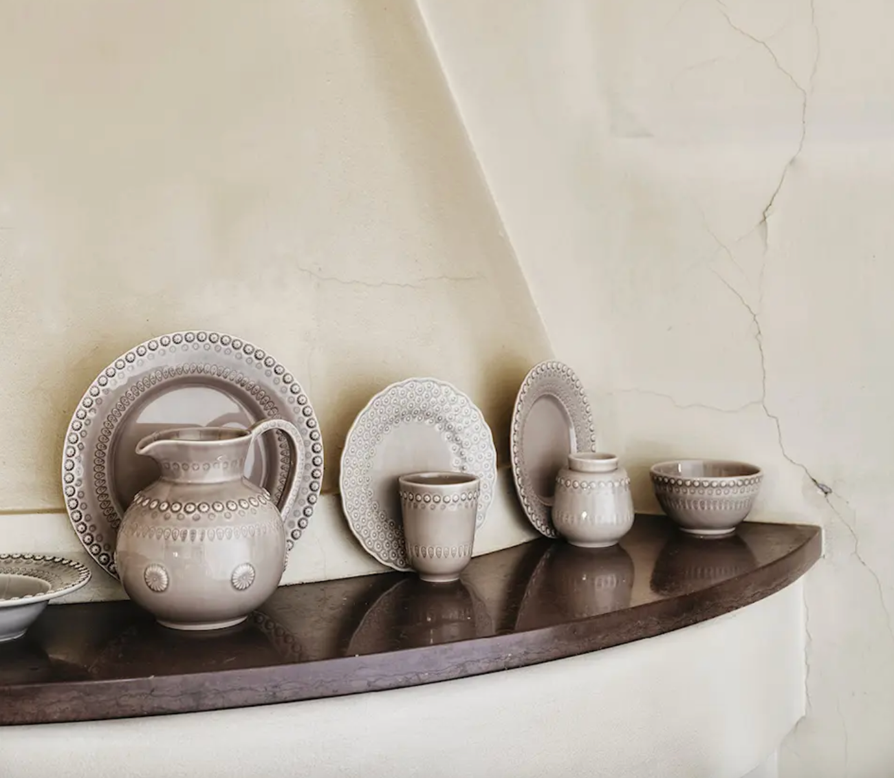 PotteryJo Daisy servis i beige stengods – tallrikar, skålar, kanna och muggar med präglad blommönstrad kant, uppställda mot rustik vägg på mörk hylla.
