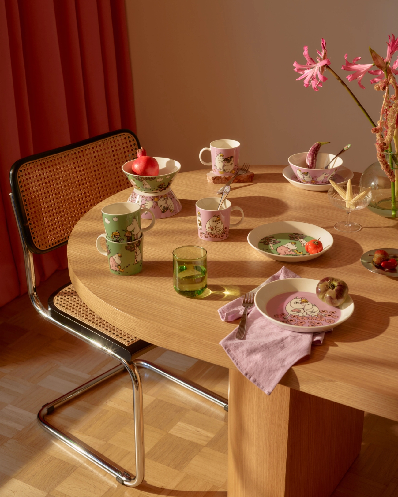 A wooden dining table is set with pink and green Moomin-themed tableware, illuminated by warm sunlight.