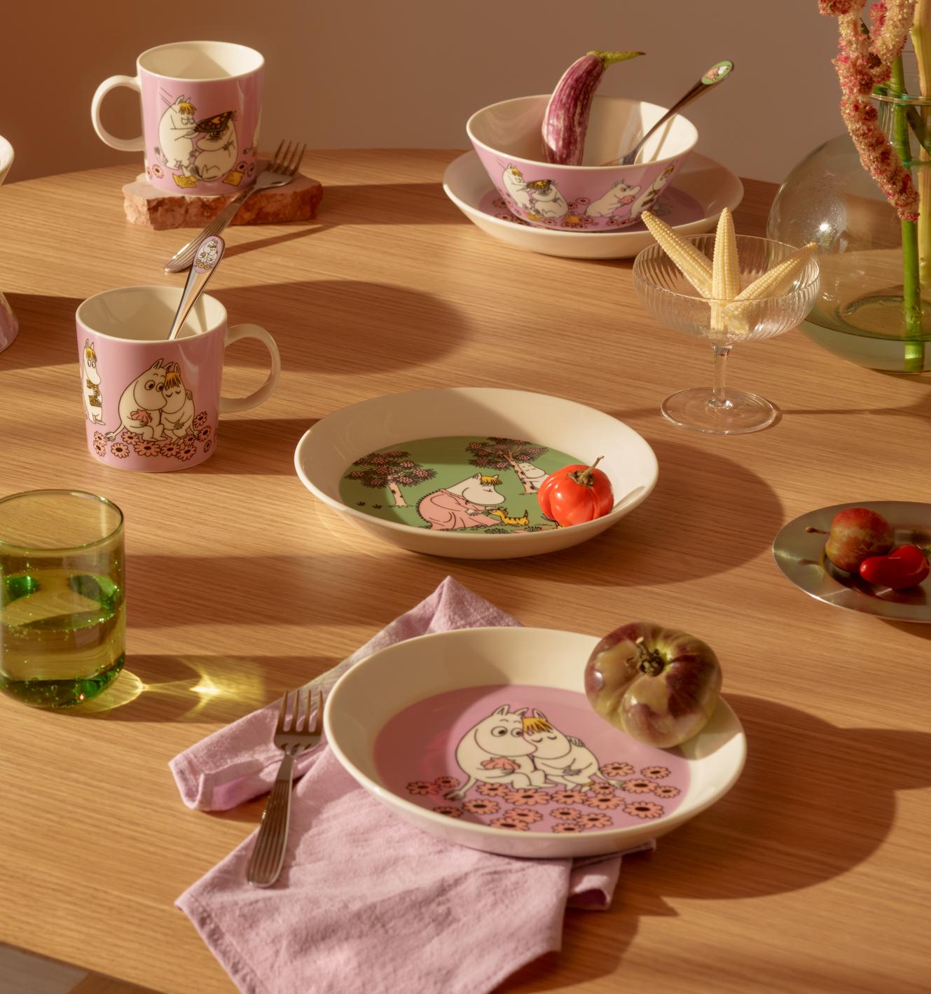 Ett matbord i trä dukat med rosa och grönt Mumin-porslin, färsk frukt och blommor, badande i solljus.