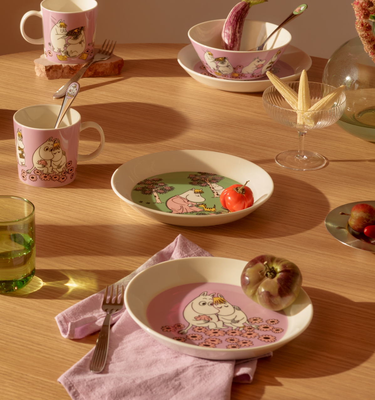 Ett matbord i trä dukat med rosa och grönt Mumin-porslin, färsk frukt och blommor, badande i solljus.