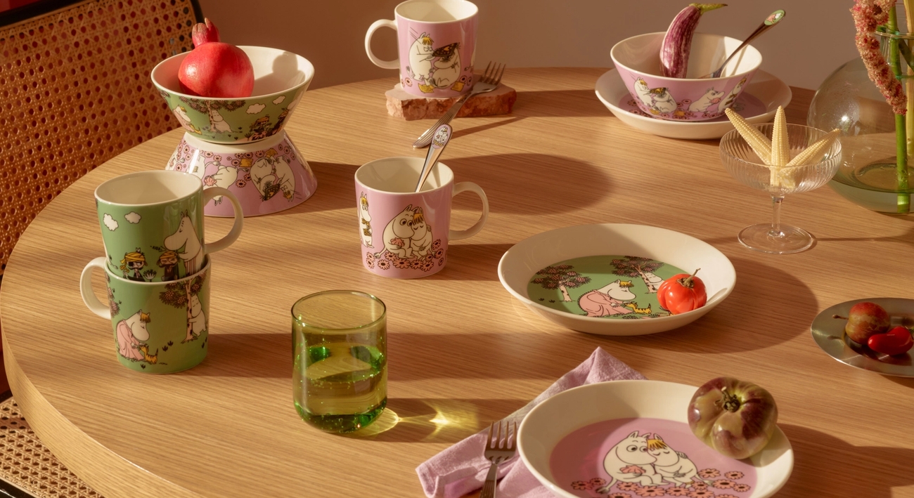 Ett matbord i ljust trä med rosa och grönt Muminporslin, frukter och blommor, badande i solljus.