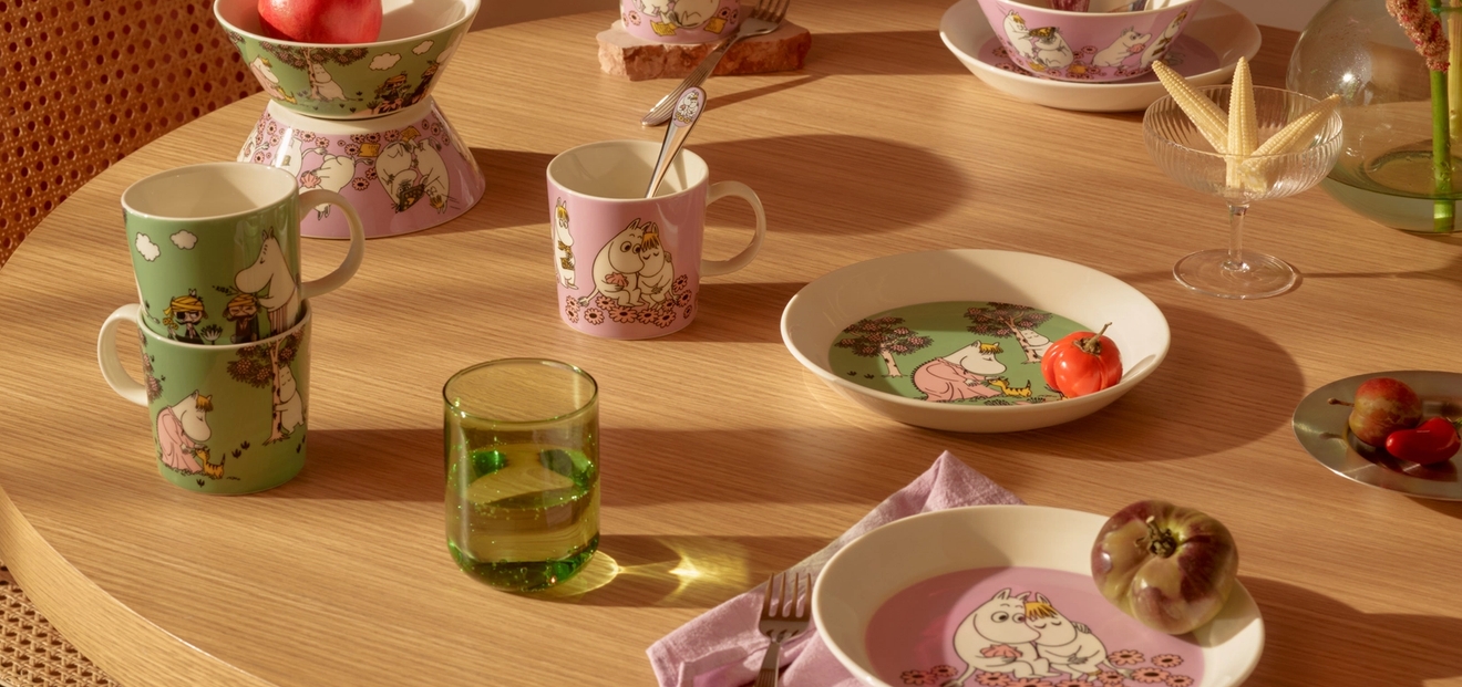 Ett matbord i ljust trä med rosa och grönt Muminporslin, frukter och blommor, badande i solljus.