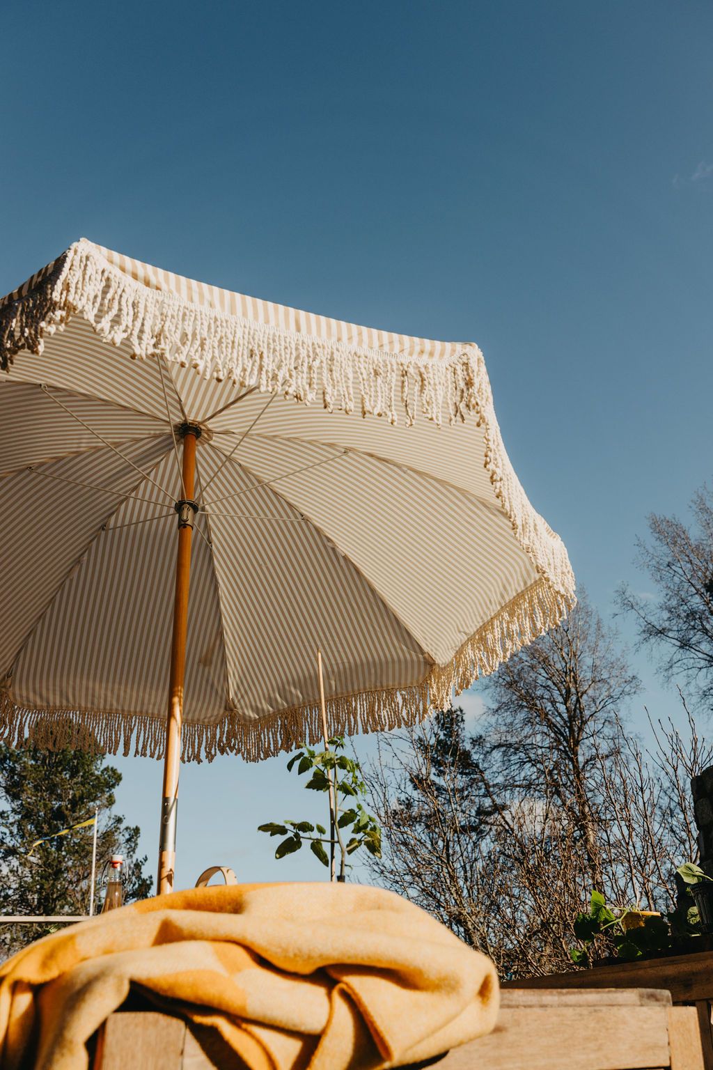 En stripete beige og hvit parasoll med frynser under en blå himmel, med et gult teppe på et bord
