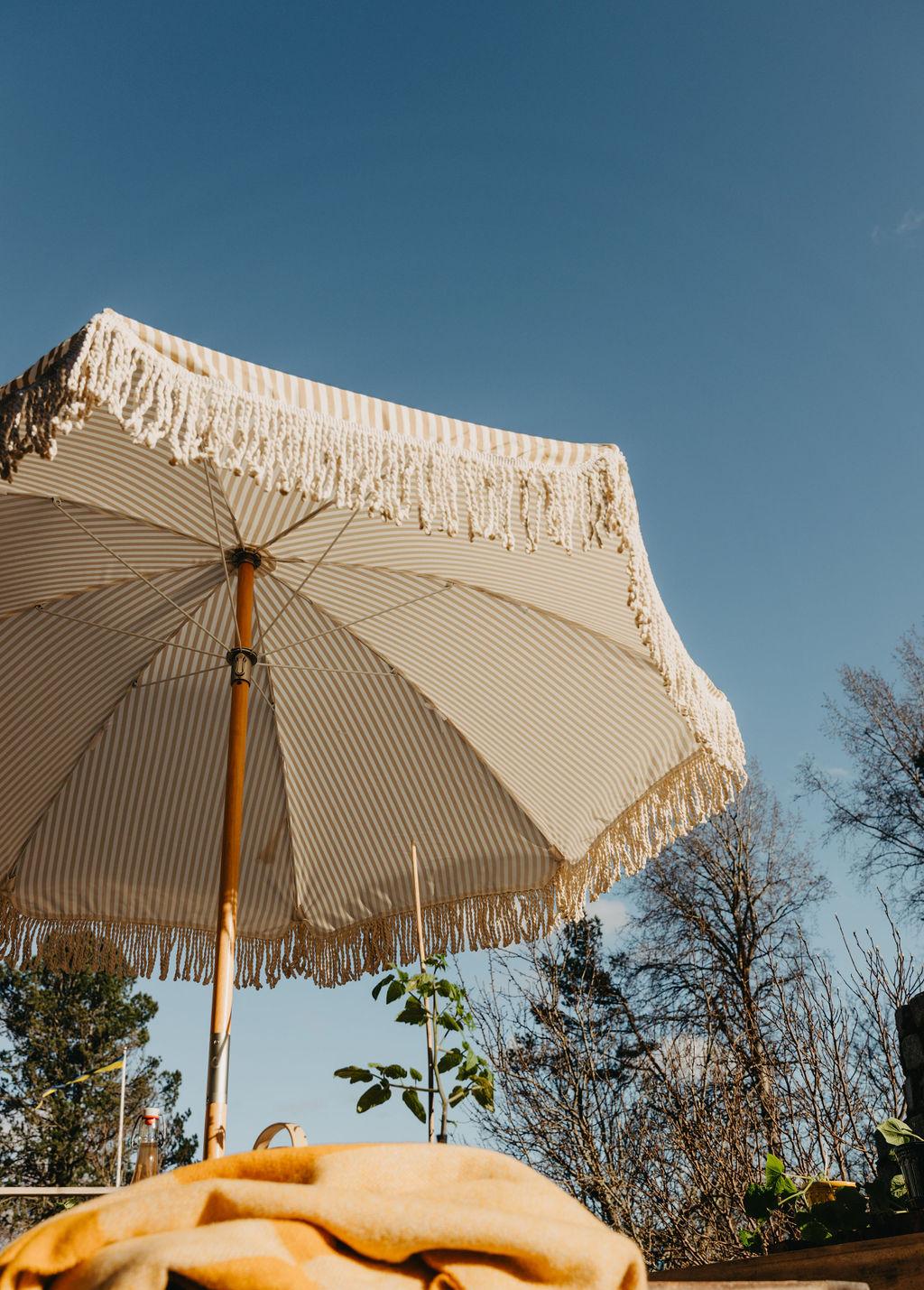 Raidallinen beige-valkoinen aurinkovarjo hapsuilla sinisen taivaan alla, sekä keltainen viltti pöydällä