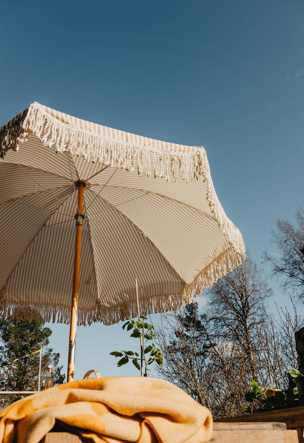 En stripete beige og hvit parasoll med frynser under en blå himmel, med et gult teppe på et bord