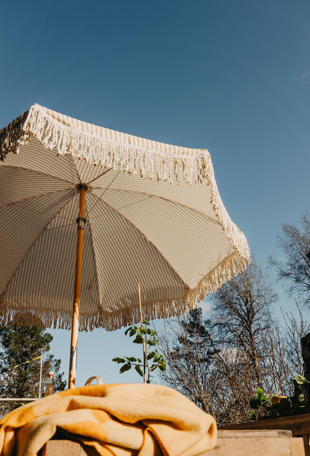 Ett randigt beige- och vitfärgat parasoll med fransar under en blå himmel, med en gul filt på ett bord