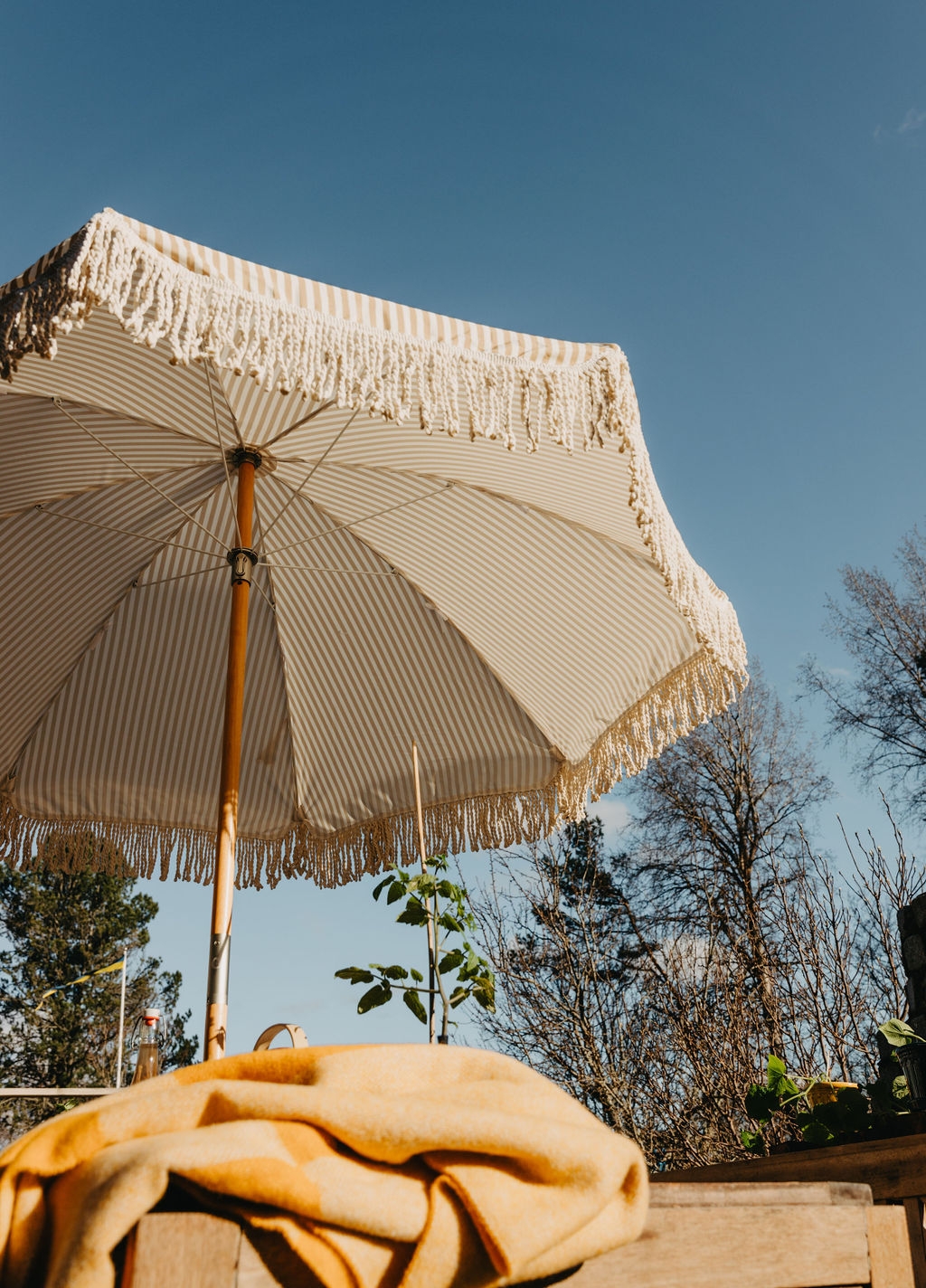 En stripete beige og hvit parasoll med frynser under en blå himmel, med et gult teppe på et bord