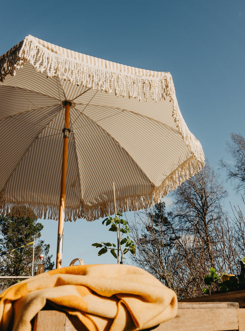 Ett randigt beige- och vitfärgat parasoll med fransar under en blå himmel, med en gul filt på ett bord