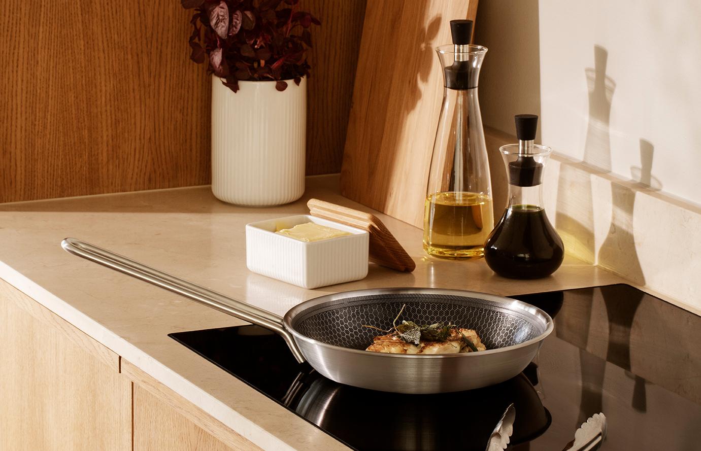 En modern köksbänkskiva med mat som tillagas på induktionshäll, en smörform, en krukväxt och trädetaljer.