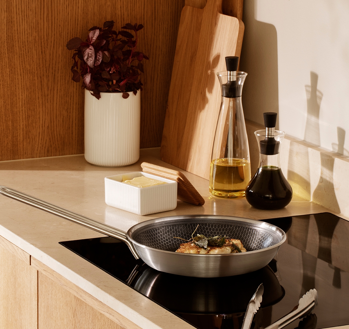 Ruokaa kypsennetään pannulla induktioliedellä kahden öljyannostelijan ja puisten leikkuulautojen kanssa aurinkoisessa keittiössä.