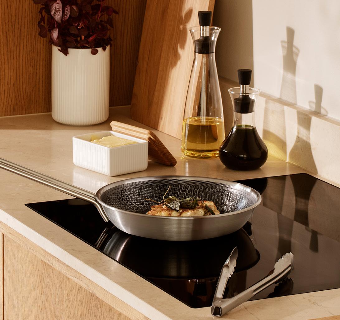 Moderni keittiötaso, jonka paistinpannulla kypsennetään ruokaa induktioliedellä, ja jota reunustavat öljypullot, voivuoka ja puiset leikkuulaudat.