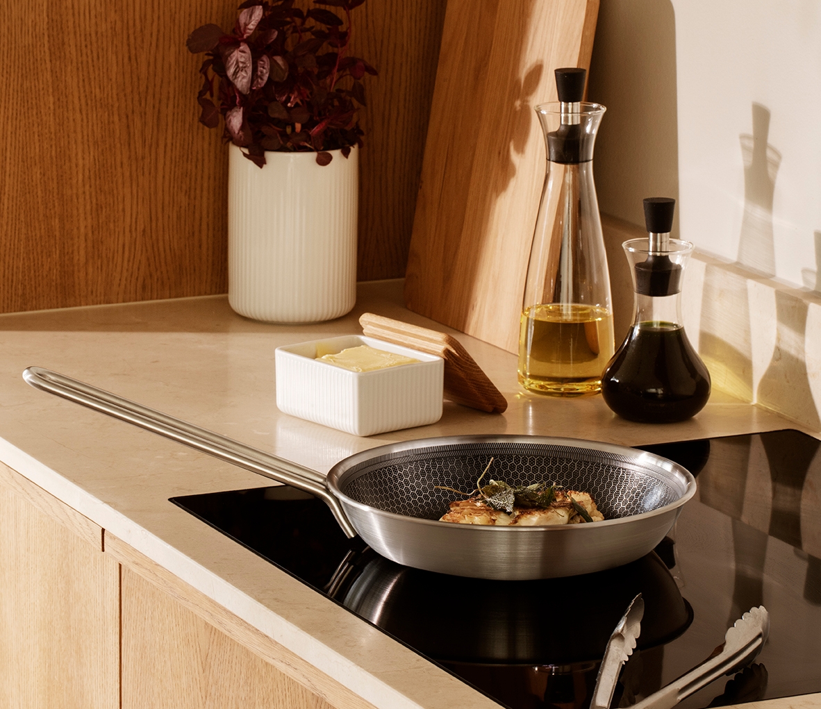 En modern köksbänkskiva med mat som tillagas på induktionshäll, en smörform, en krukväxt och trädetaljer.