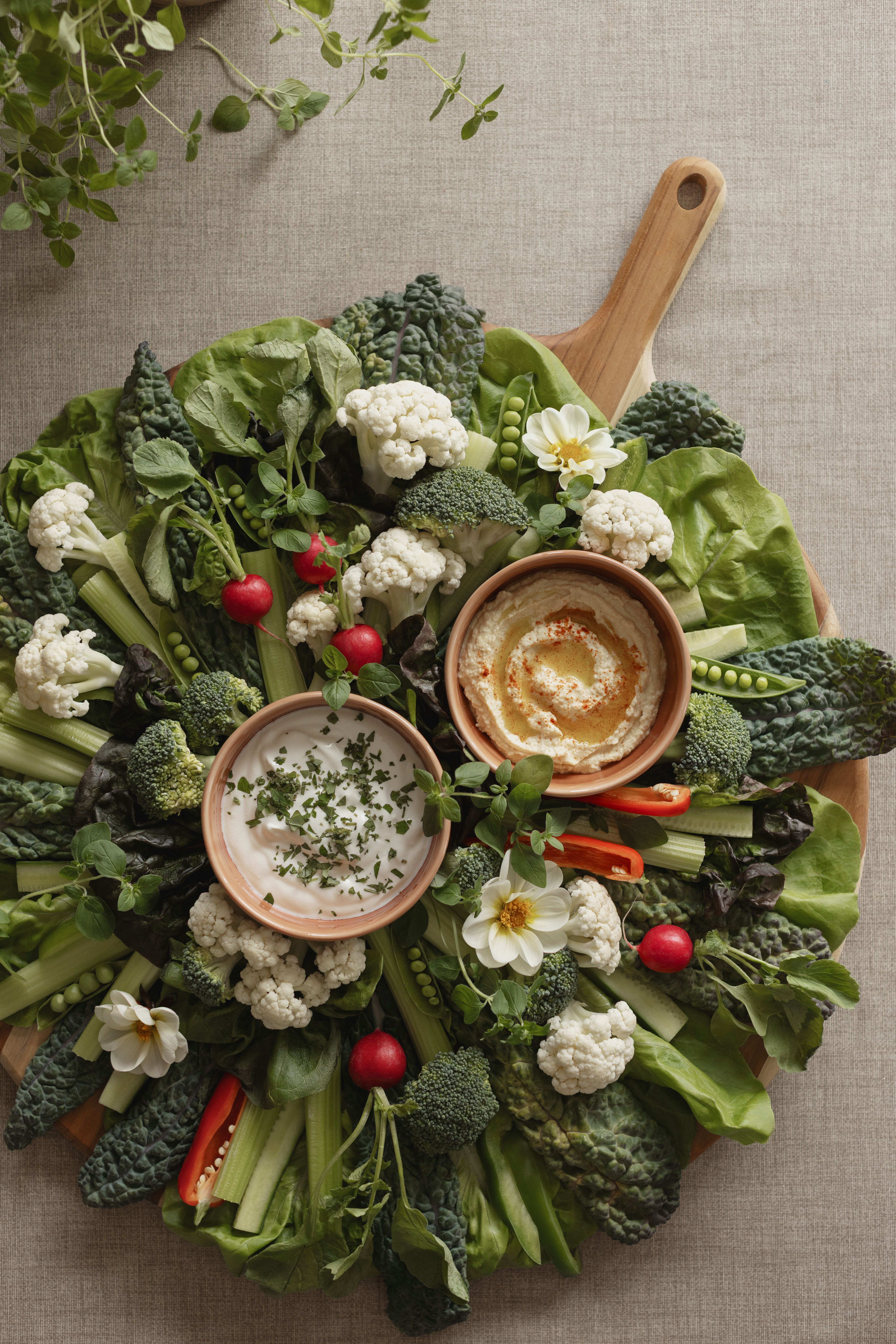 Färgglad grönsaksbricka med blomkål, broccoli, rädisor, selleri och sallad runt två skålar med dippsåser.