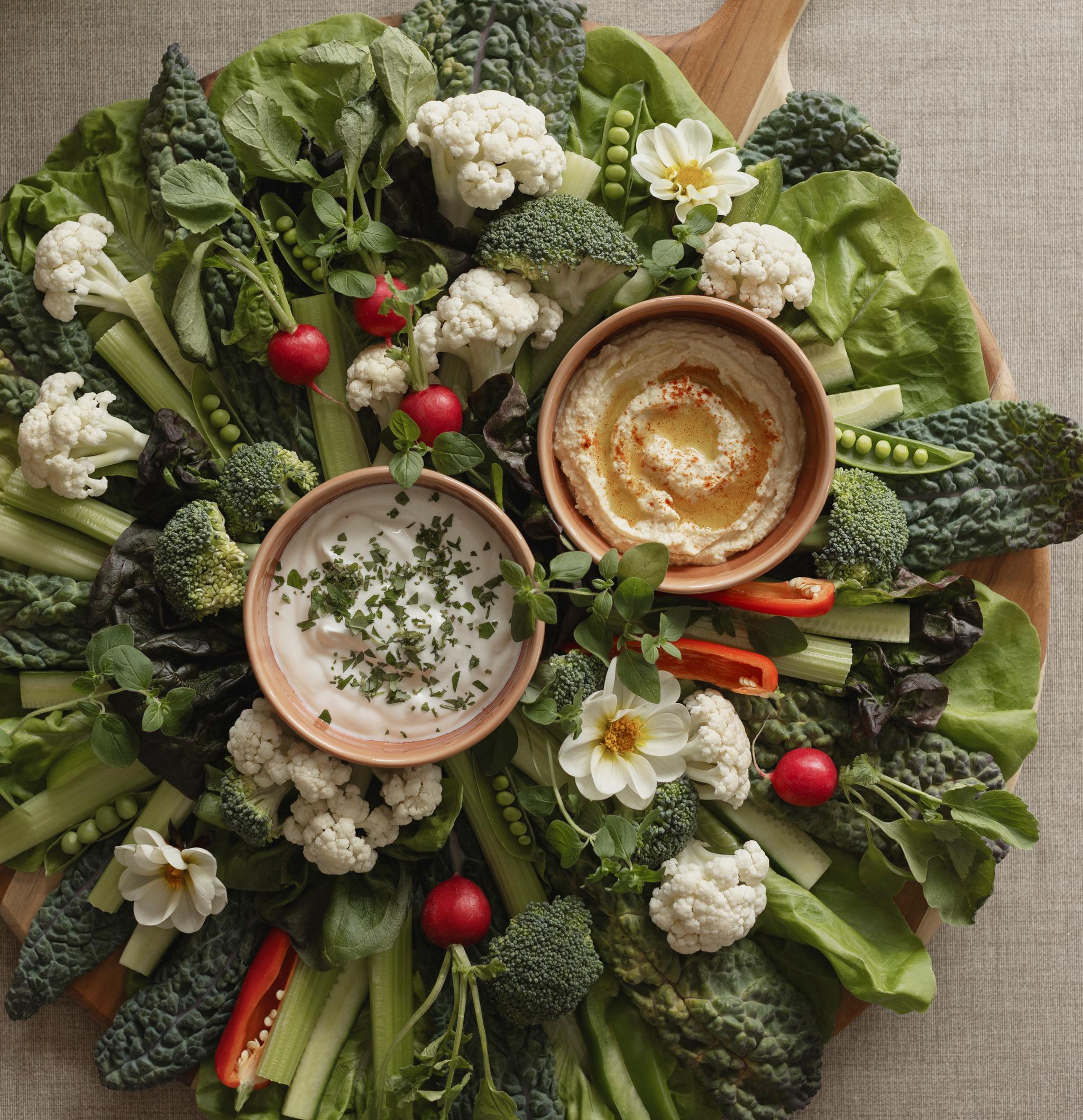 Färgglad grönsaksbricka med blomkål, broccoli, rädisor, selleri och sallad runt två skålar med dippsåser.