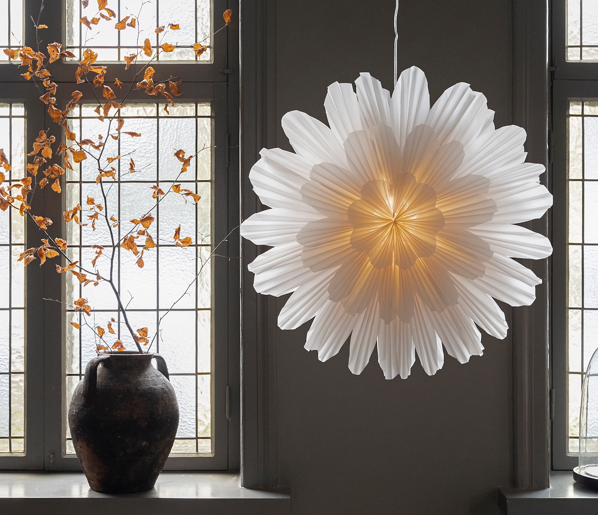 Stor vit blomformad papperslampa lyser framför spröjsade fönster bredvid en mörk vas med höstgrenar.