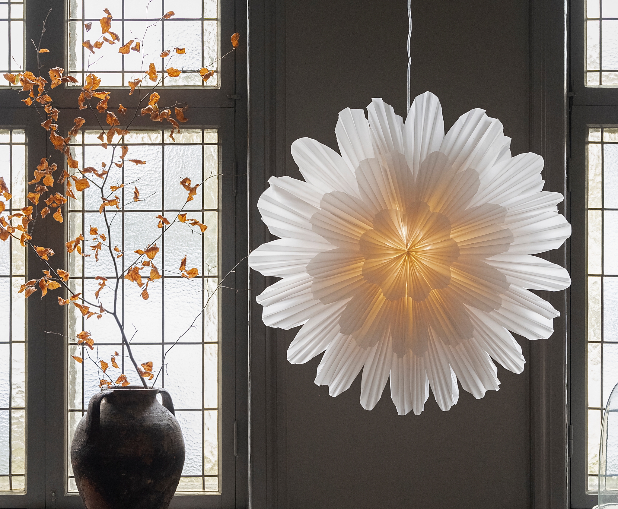Stor vit blomformad papperslampa lyser framför spröjsade fönster bredvid en mörk vas med höstgrenar.