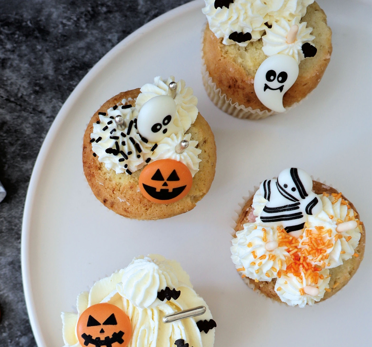Tre Halloween-cupcakes dekorerade med pumpor, fladdermöss och en mumie ligger på en vit tallrik bredvid tuber med strössel.