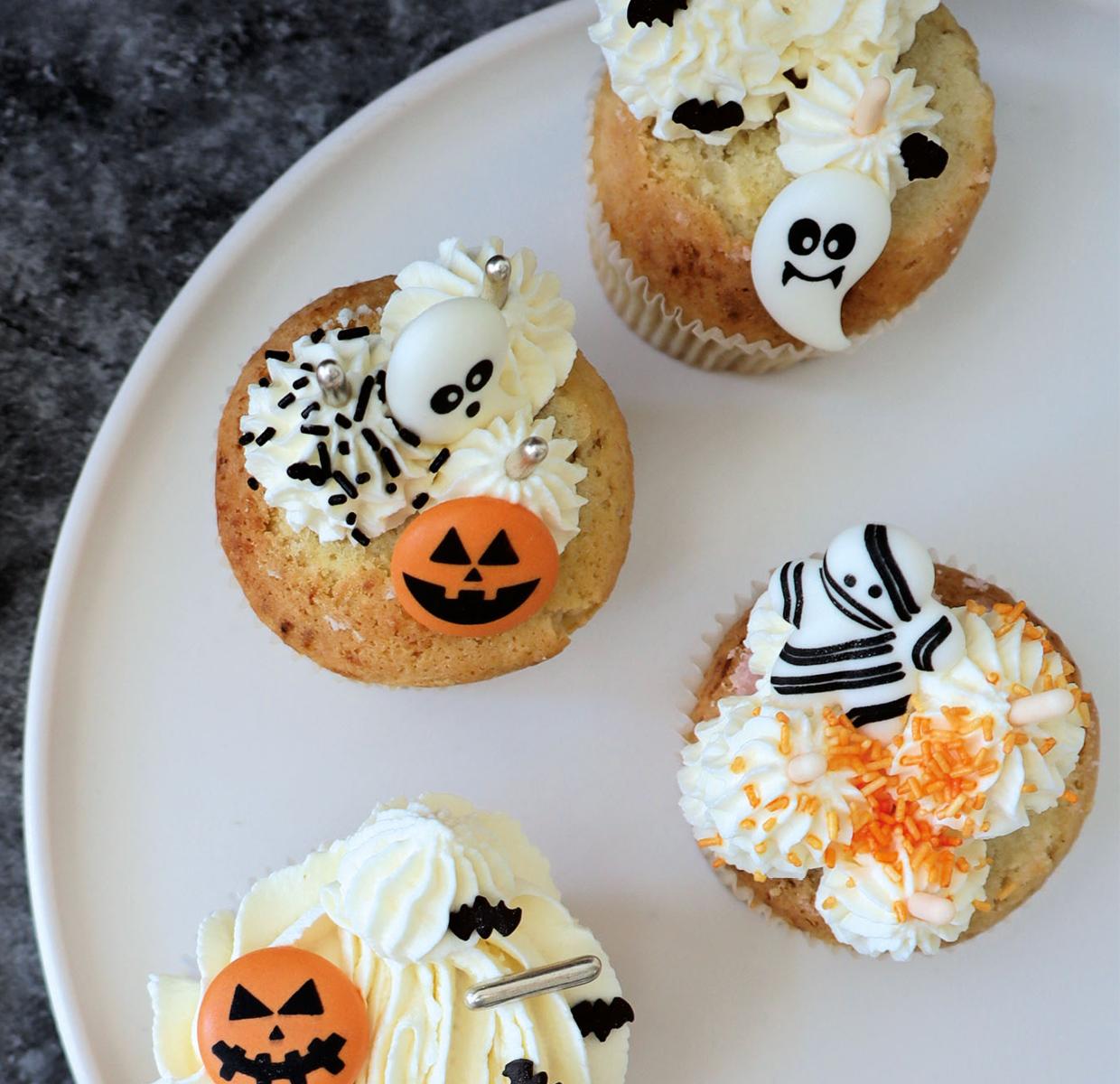 Tre Halloween-cupcakes dekorerade med pumpor, fladdermöss och en mumie ligger på en vit tallrik bredvid tuber med strössel.