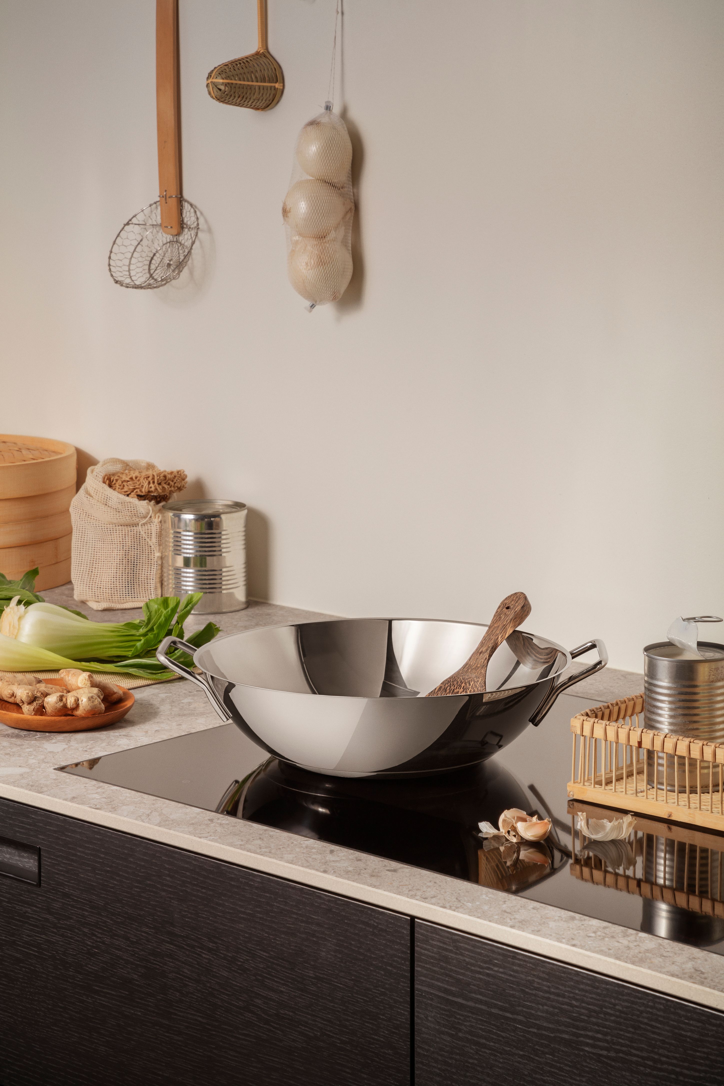 En wok i rustfritt stål med en treskje står på en induksjonstopp, omgitt av ingefær, bok choy og hvitløk på en kjøkkenbenk.